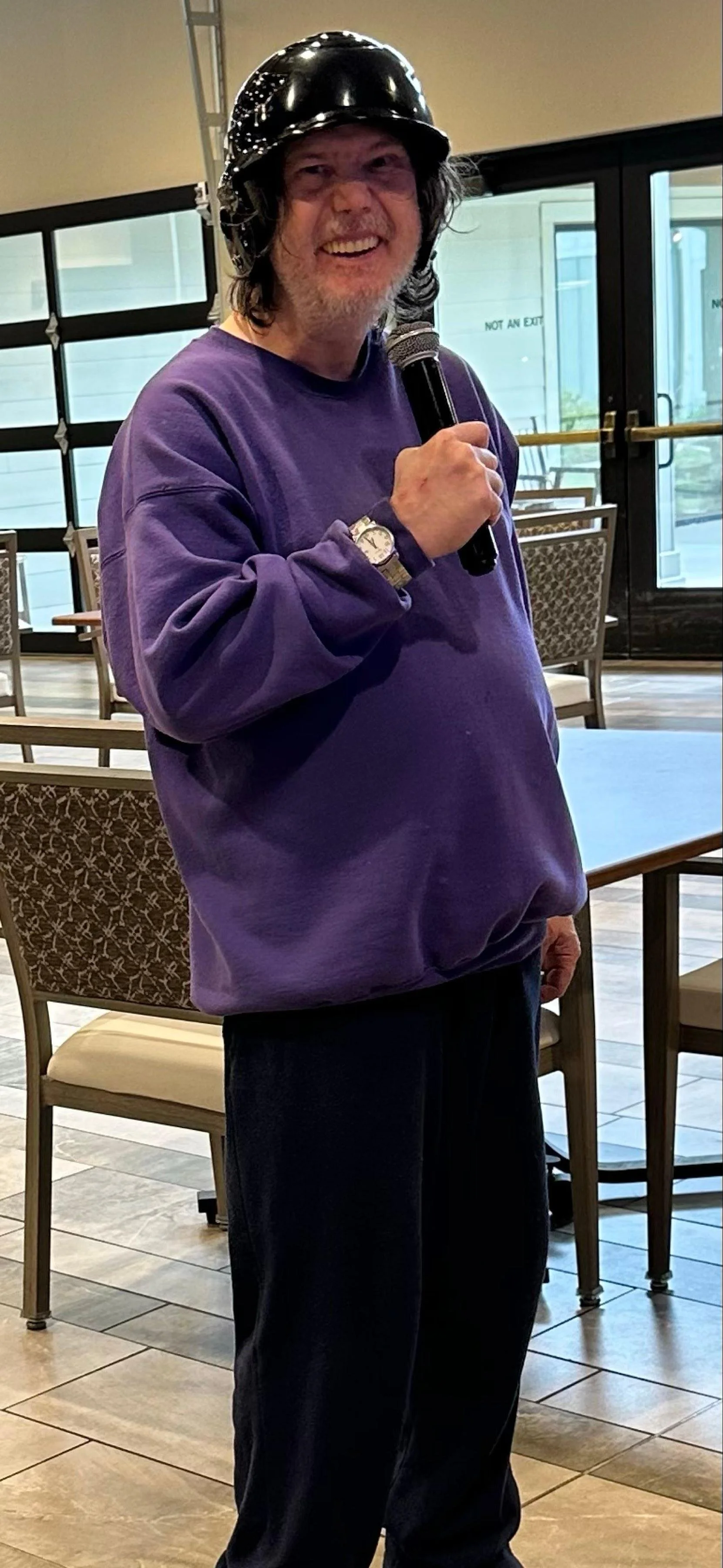 A man with long hair and a beard wearing a black helmet, a purple sweatshirt, and dark pants, holding a microphone and smiling indoors with chairs and windows in the background.