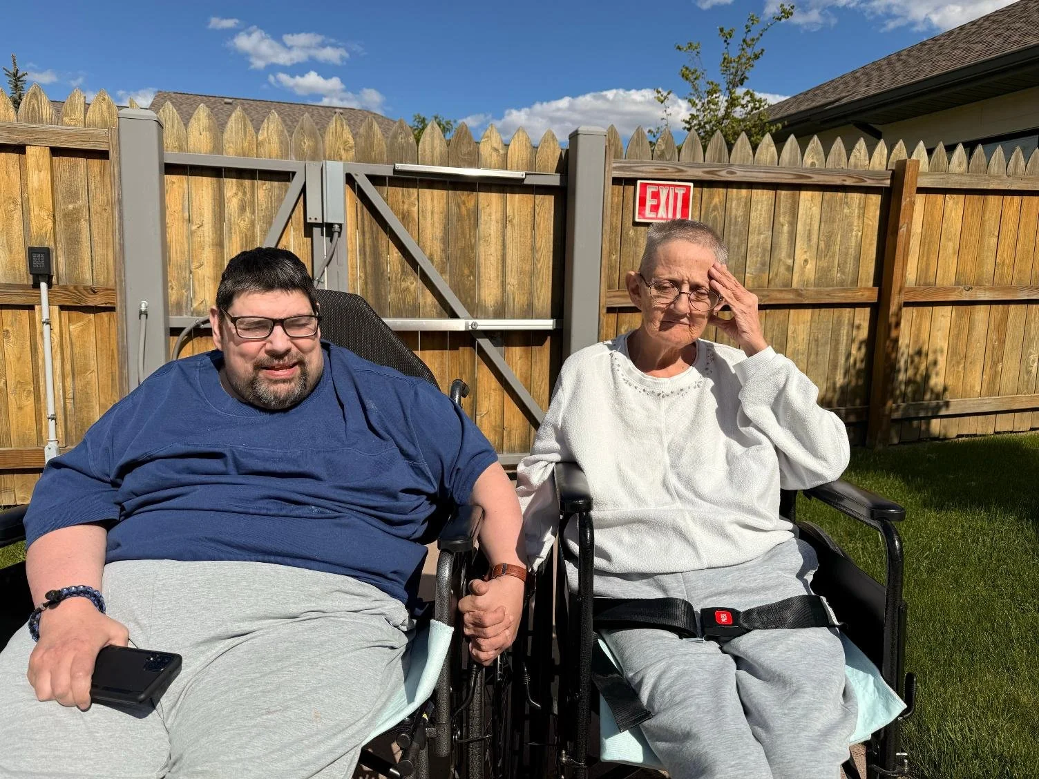 A man and an elderly woman sitting outside in wheelchairs, holding hands, with a wooden fence and blue sky in the background.