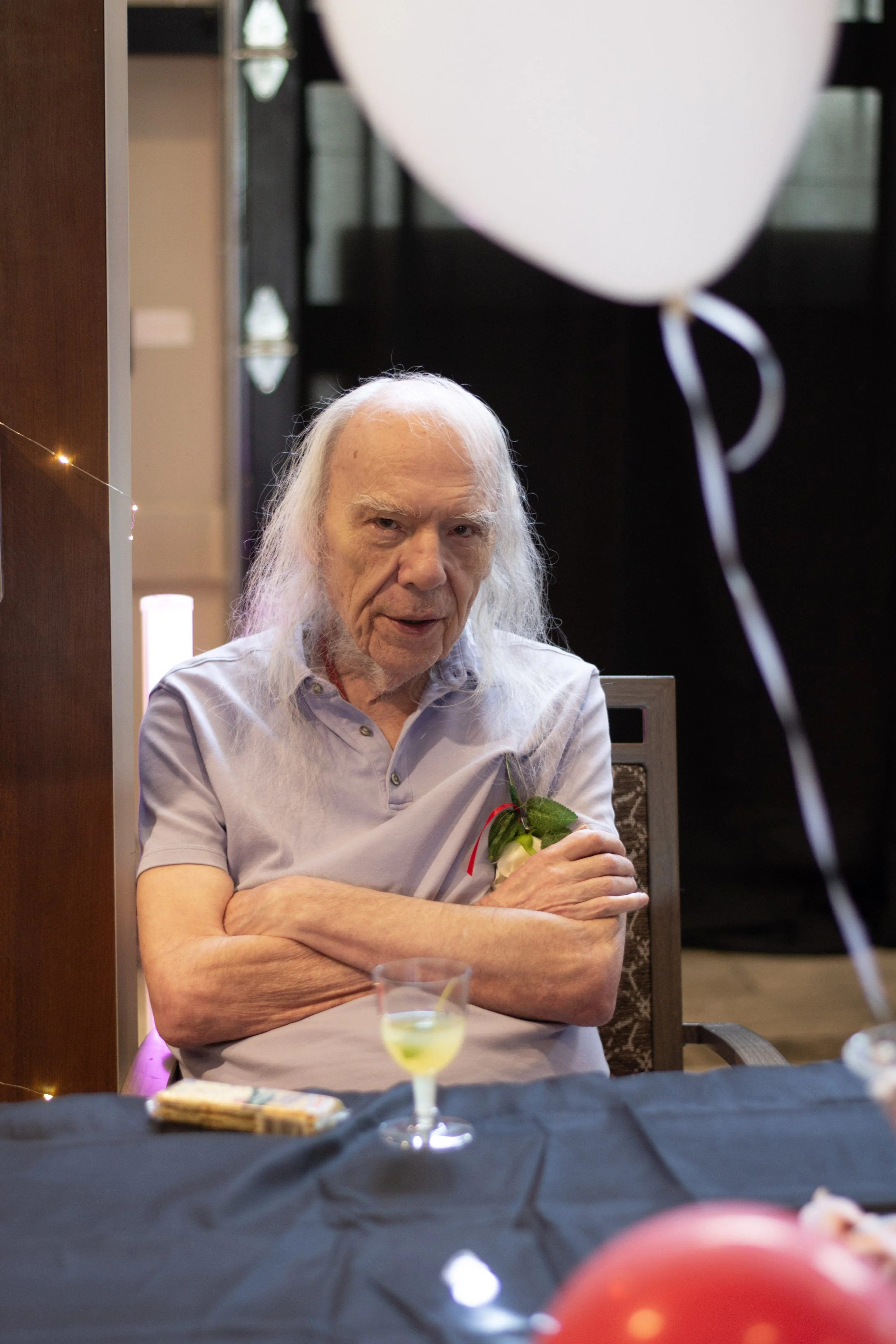 Older man with long white hair and a mustache sitting at a table with a black tablecloth, holding a flower with a small ribbon. There is a partially visible drink in front of him. A large white balloon is in the upper right corner. The background inc