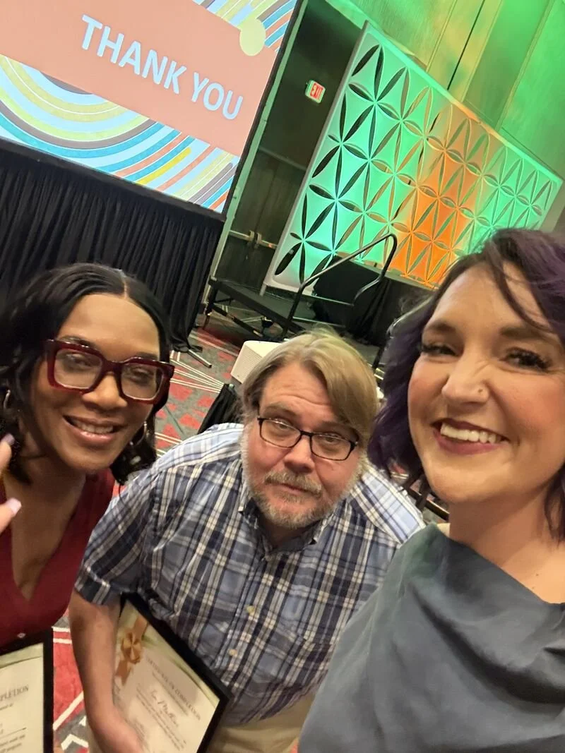 Three people taking a selfie at an indoor event, with a large screen displaying a colorful thank you message in the background.