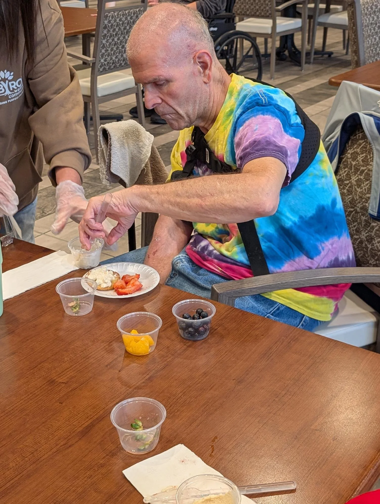 A man with a shaved head wearing a colorful tie-dye T-shirt sits at a table, reaching for food in a small clear cup. The table has various small containers with different types of fruit and food. A person next to him, wearing gloves and a brown shirt