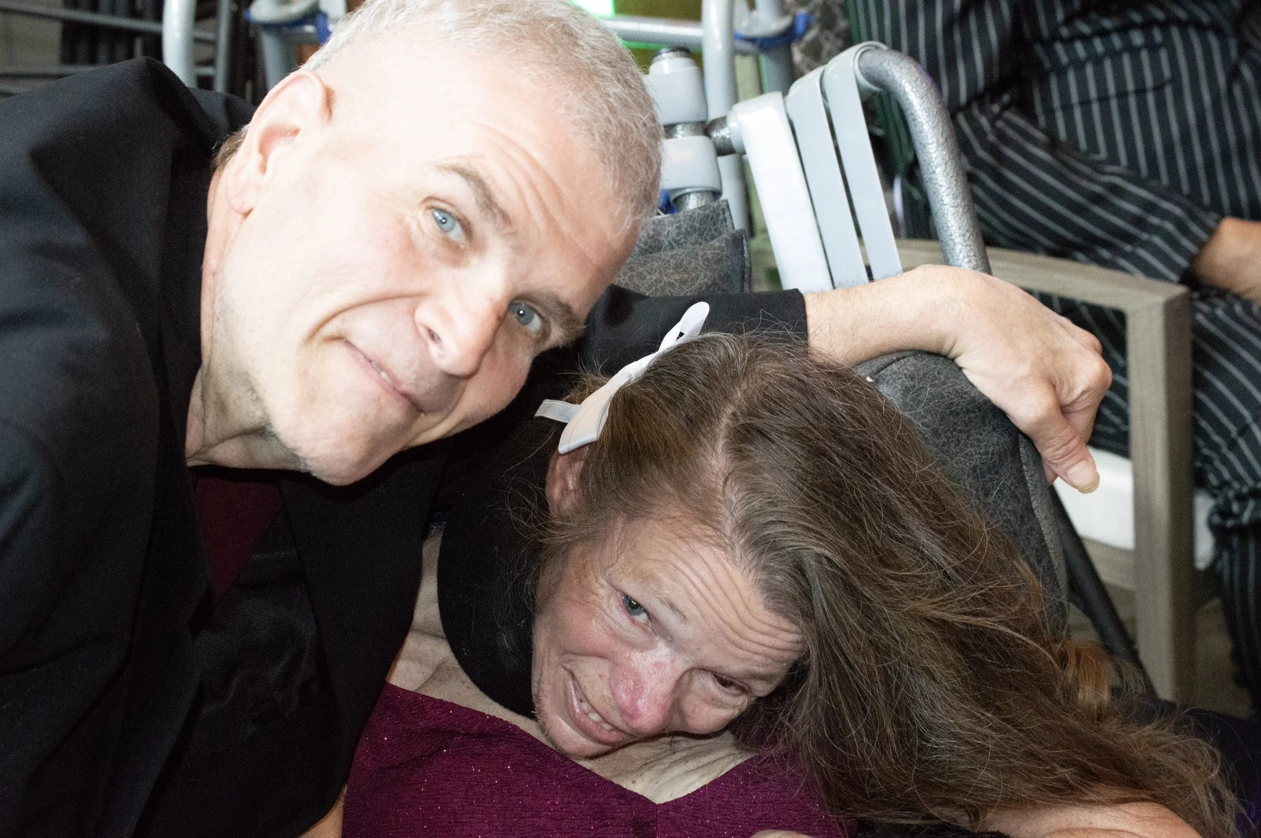 Two people lying on the floor with their heads close together. One woman with long brown hair and glasses, and a man with short blonde hair and blue eyes in the background. They are smiling and appear to be resting or relaxing.