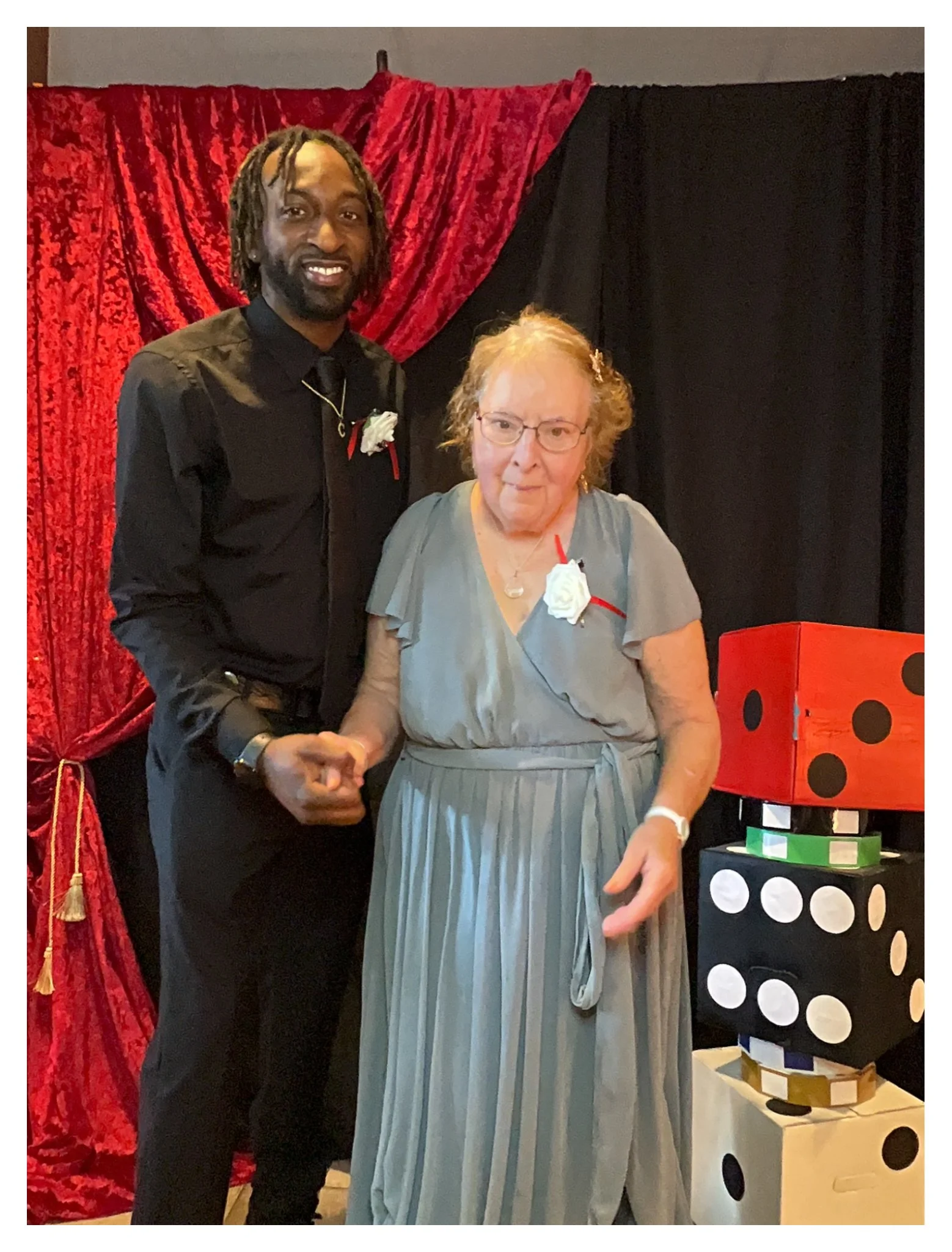 A man and a woman shaking hands at a celebration or party. The man is wearing a black shirt with a white boutonnière and a necklace, while the woman is wearing a light gray dress with a white rose corsage. The background has red and black curtains, a