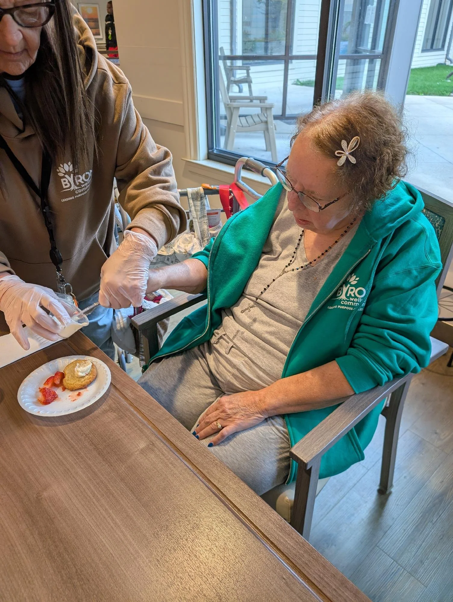 A woman sitting in a chair with a teal jacket and gray shirt, receiving an injection from a healthcare worker. The woman has glasses and a hair clip. The table in front of her has a plate with strawberry and a pastry. The setting is a room with large