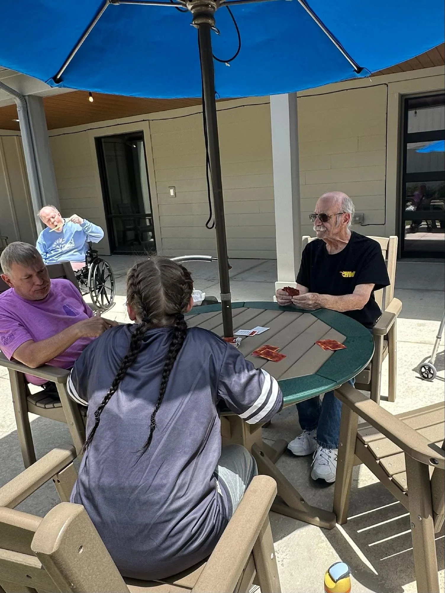 A group of elderly and middle-aged people sitting around a round outdoor table under a blue patio umbrella. One elderly woman in a wheelchair is eating with chopsticks, while a man with sunglasses and a black t-shirt is holding playing cards. Two oth