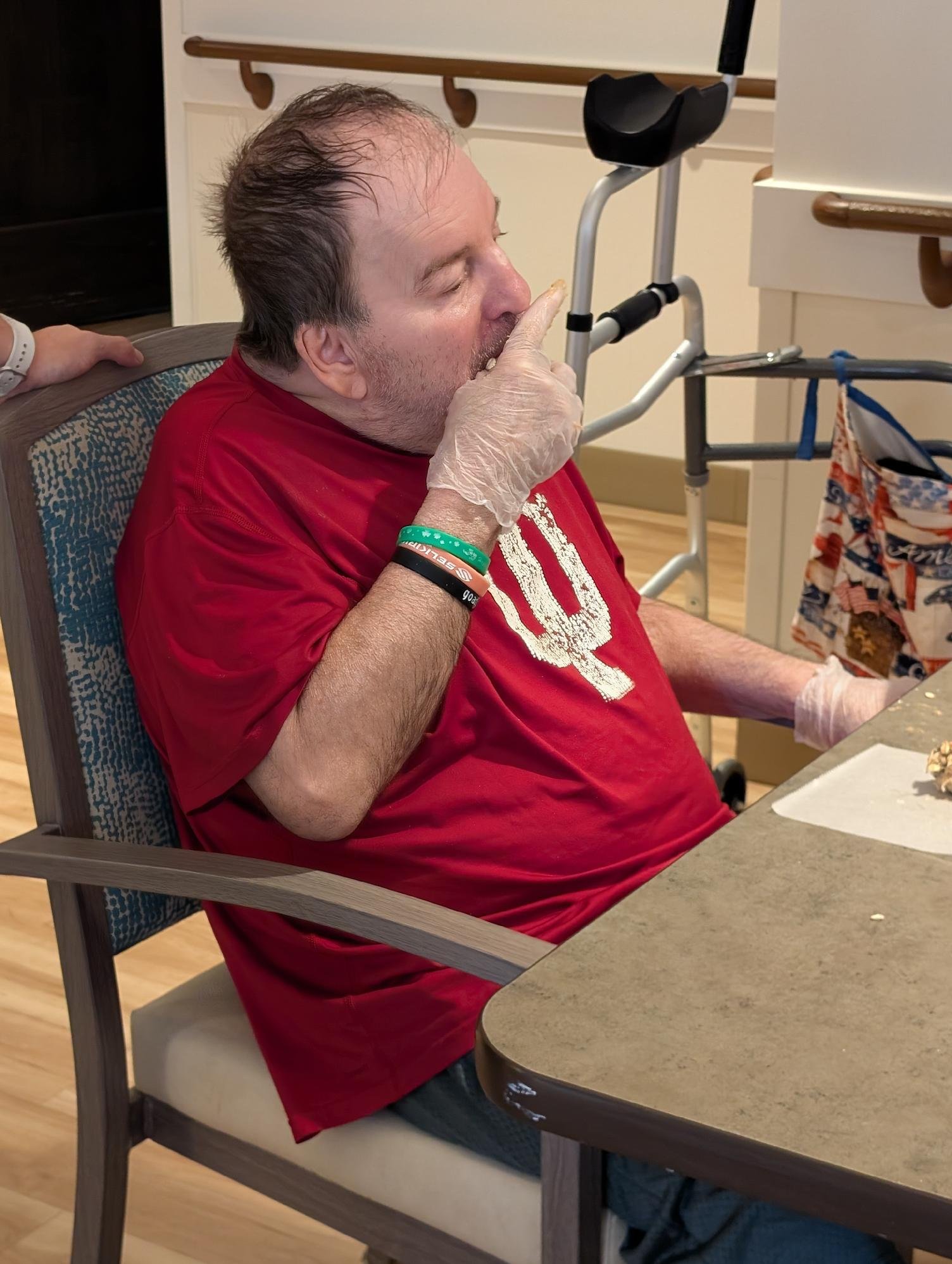 An elderly man with a band on his wrist, wearing a red shirt, sitting at a dining table with a famine, using his finger to press his lips in a silent gesture. He is in a healthcare or hospital setting with a walker visible in the background.