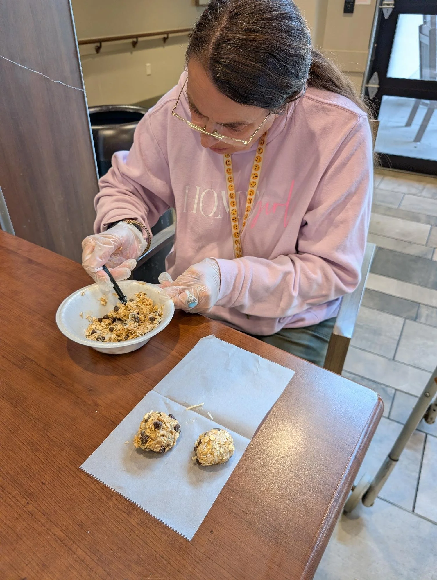 A woman wearing glasses, a pink sweatshirt with the word 'JOY' on it, and disposable gloves, is decorating oatmeal cookies with chocolate chips, raisins, and crushed nuts in a cafeteria setting.