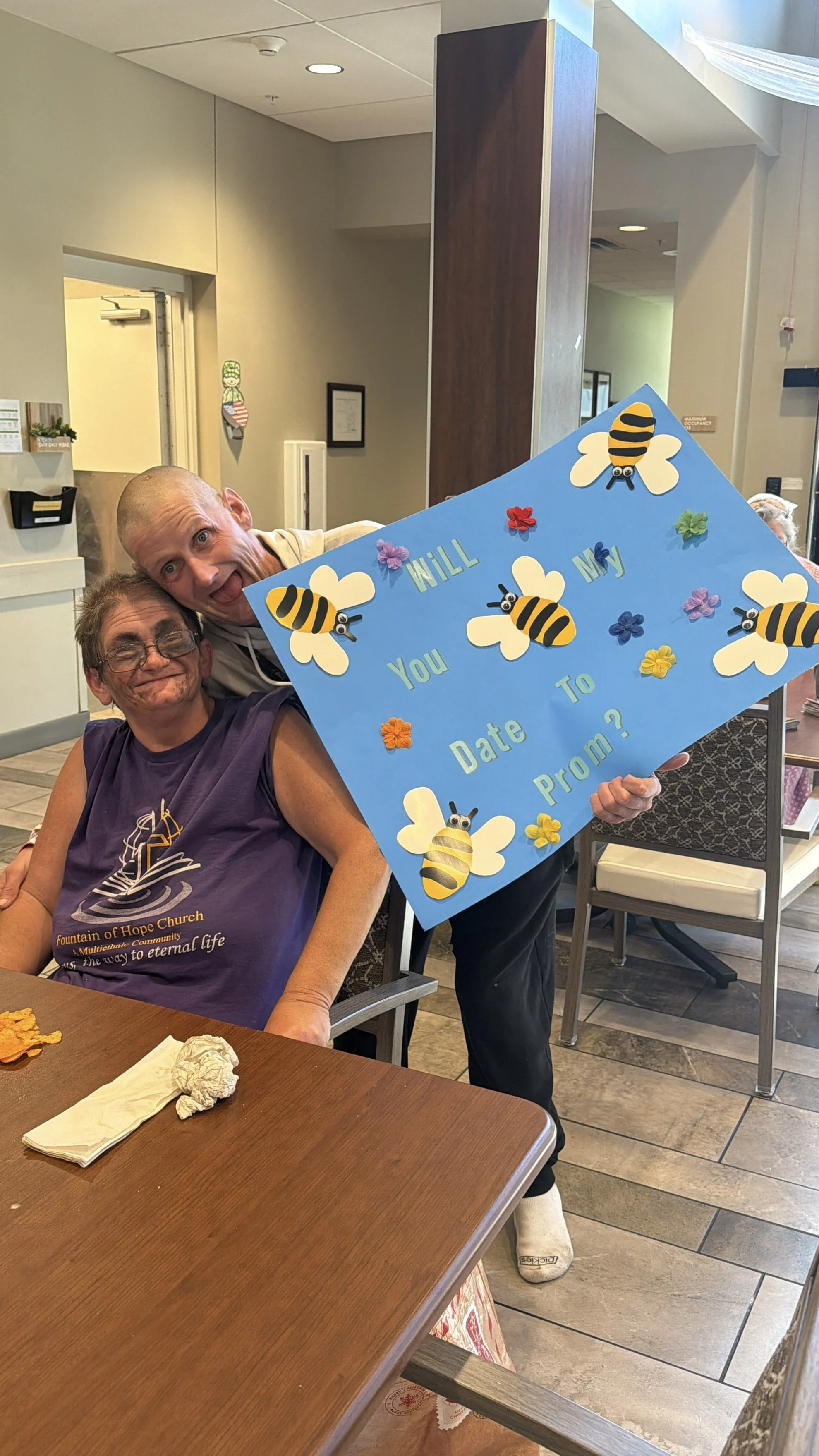 A woman is seated at a table with a comfortable smile, while another person is standing and holding a colorful sign that reads 'Will you date me to prom?' decorated with bees and flowers. The setting appears to be a restaurant or community center wit