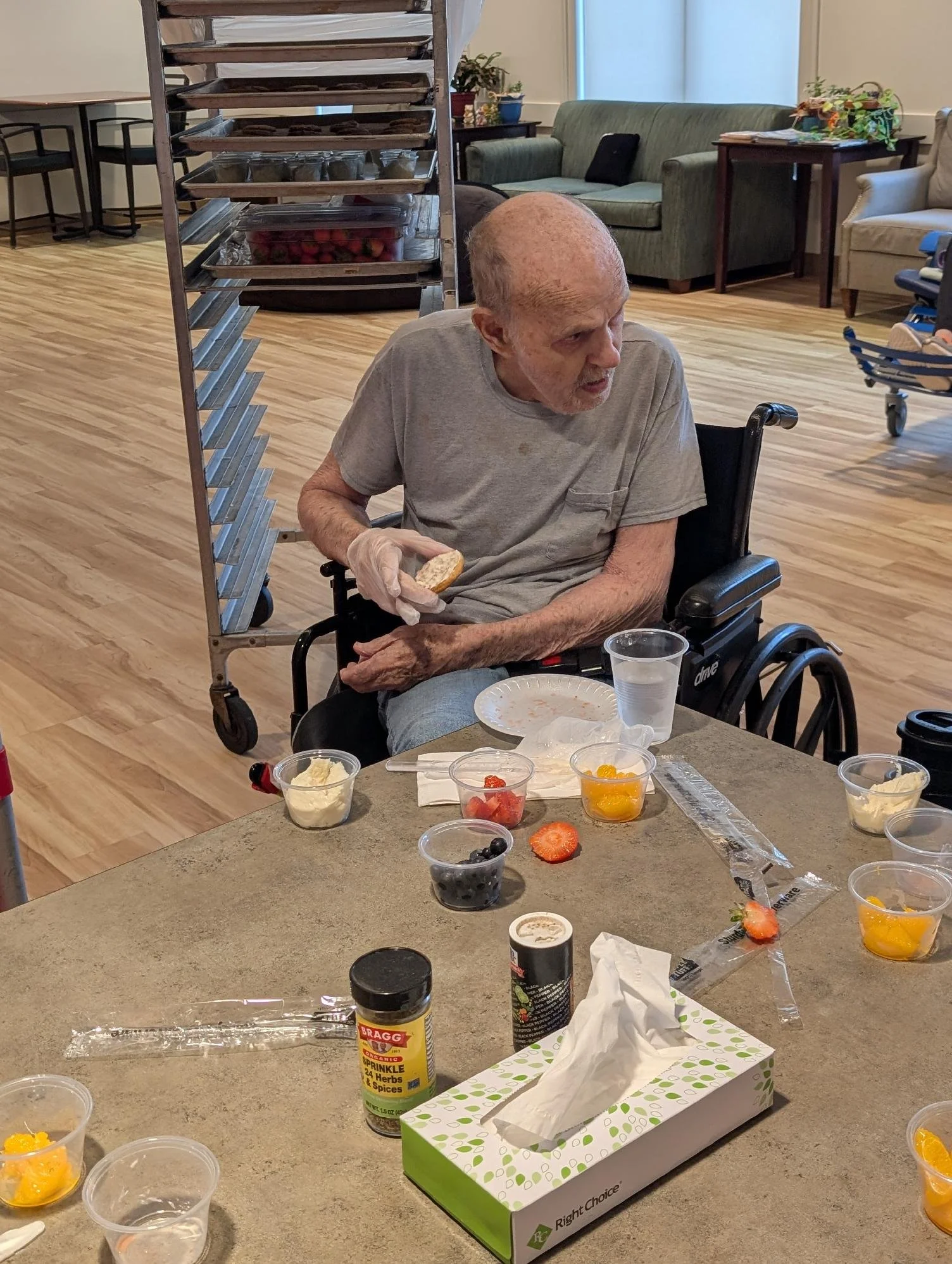 Elderly man in a wheelchair with a shaved head, wearing a gray shirt, holding a piece of bread, sitting at a table with various food items including fruit, yogurt, and containers, in a room with wooden floors and furniture.