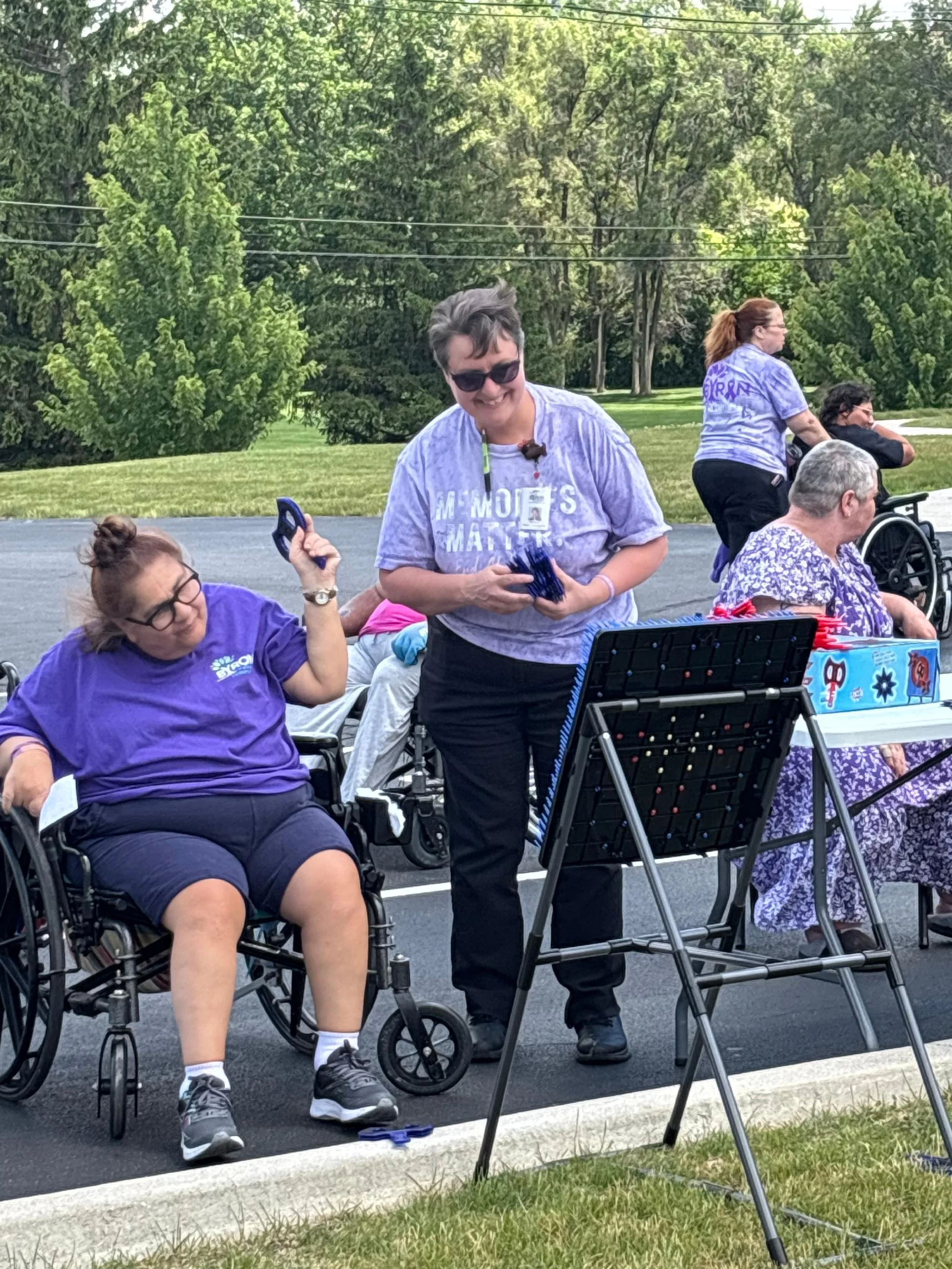 A group of people outside, some in wheelchairs, participating in an outdoor event. A woman holds a device in her hand, and others are sitting or standing. There are trees and a grassy area in the background.