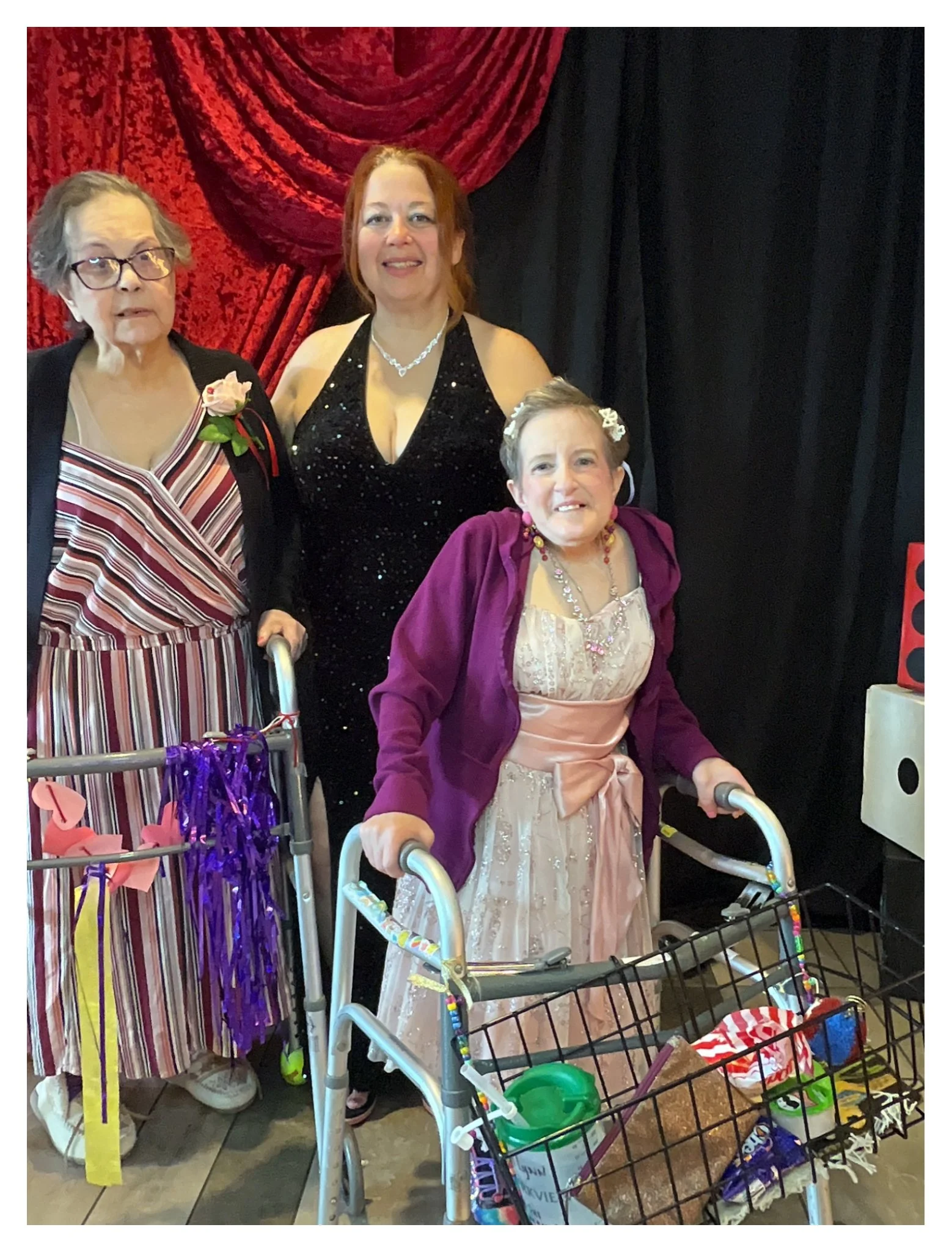 Three women at a celebration, with one woman in a pink dress in a wheelchair, another woman in a black sparkly dress, and an older woman in a striped dress and glasses, standing in front of red and black curtains, surrounded by balloons and party dec