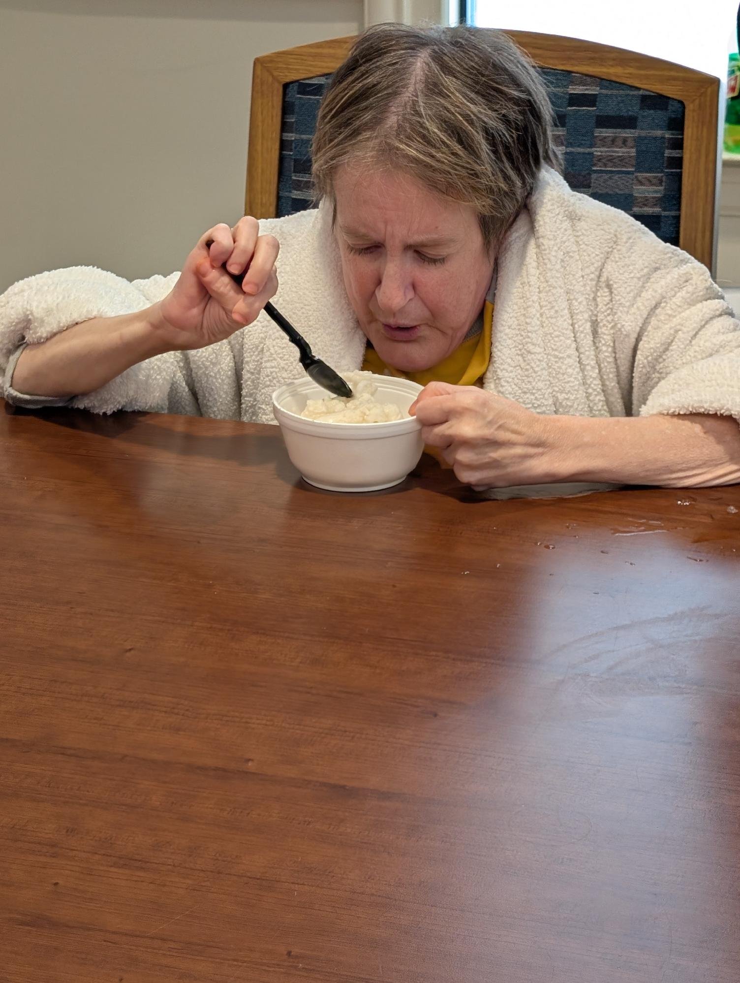 A person with short, gray hair wearing a beige fleece jacket, sitting at a wooden table, eating from a white bowl with a spoon, in a room with a chair, window, and some objects in the background.