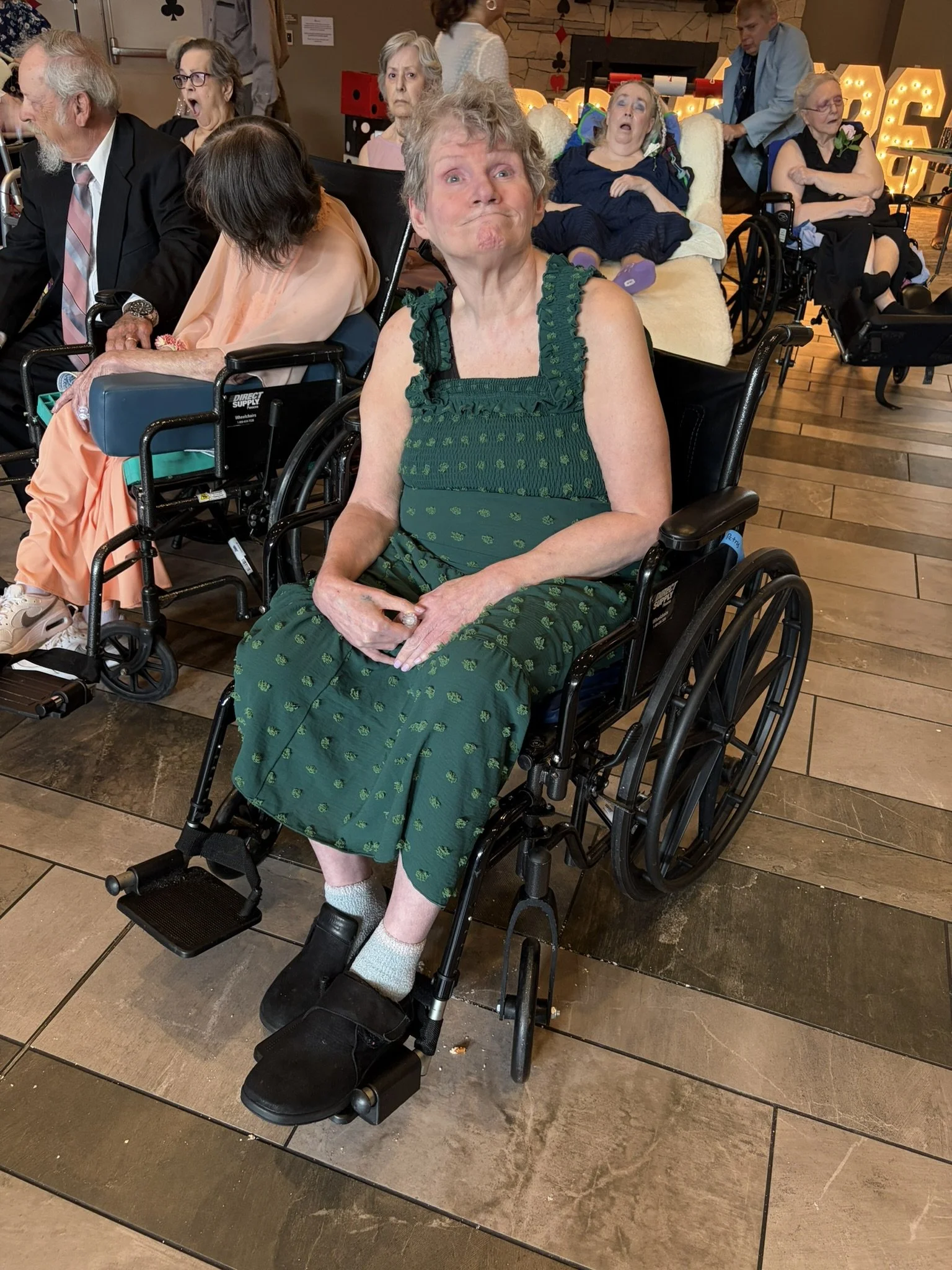 A group of elderly people sitting in wheelchairs and recliners, some with surprised or amused expressions, indoors in a room with wooden flooring and decorative lighting.