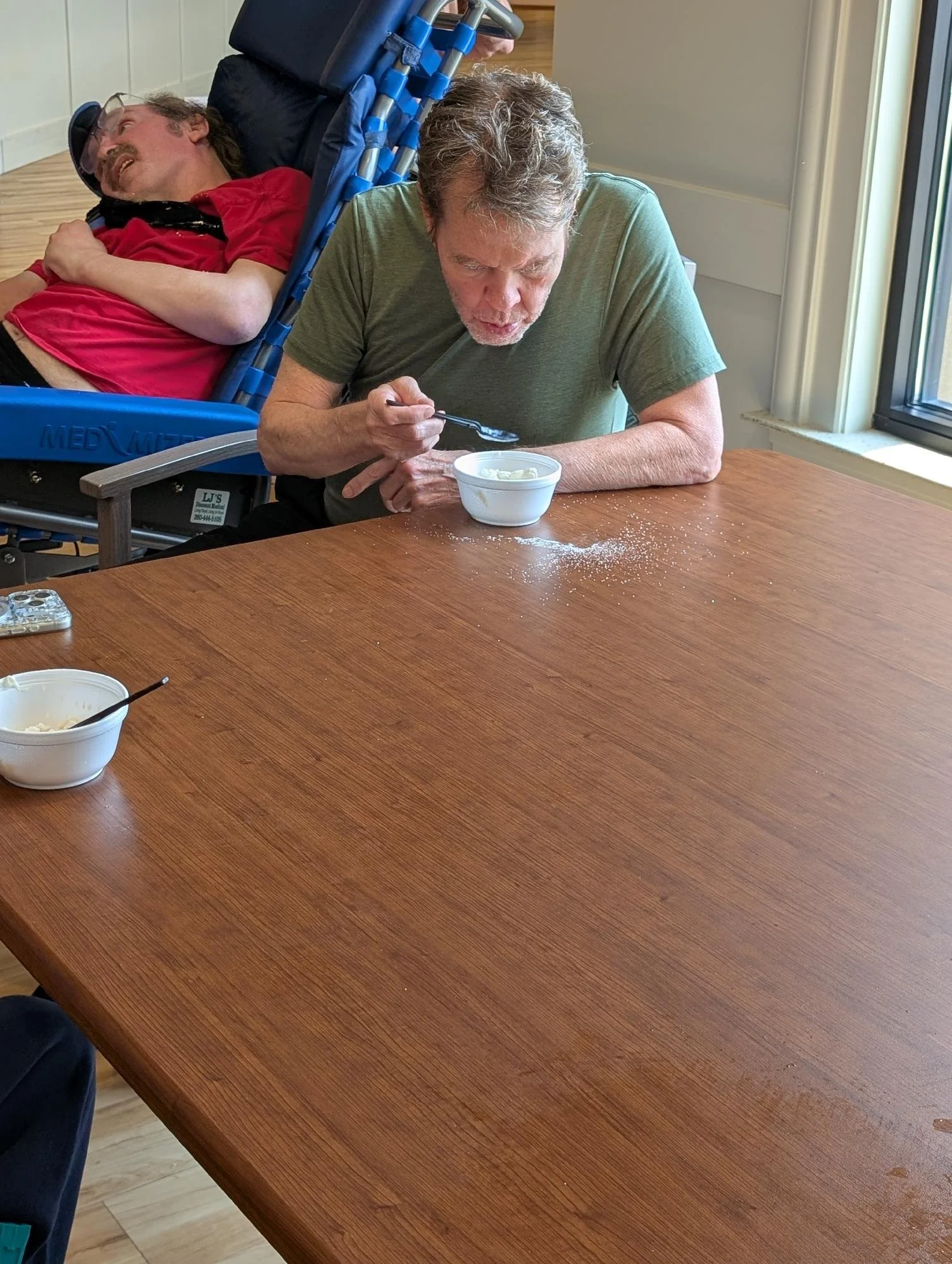 An elderly man in a green shirt eating ice cream with a spoon while sitting at a wooden table, and another man in a red shirt lying on a reclining chair in the background.