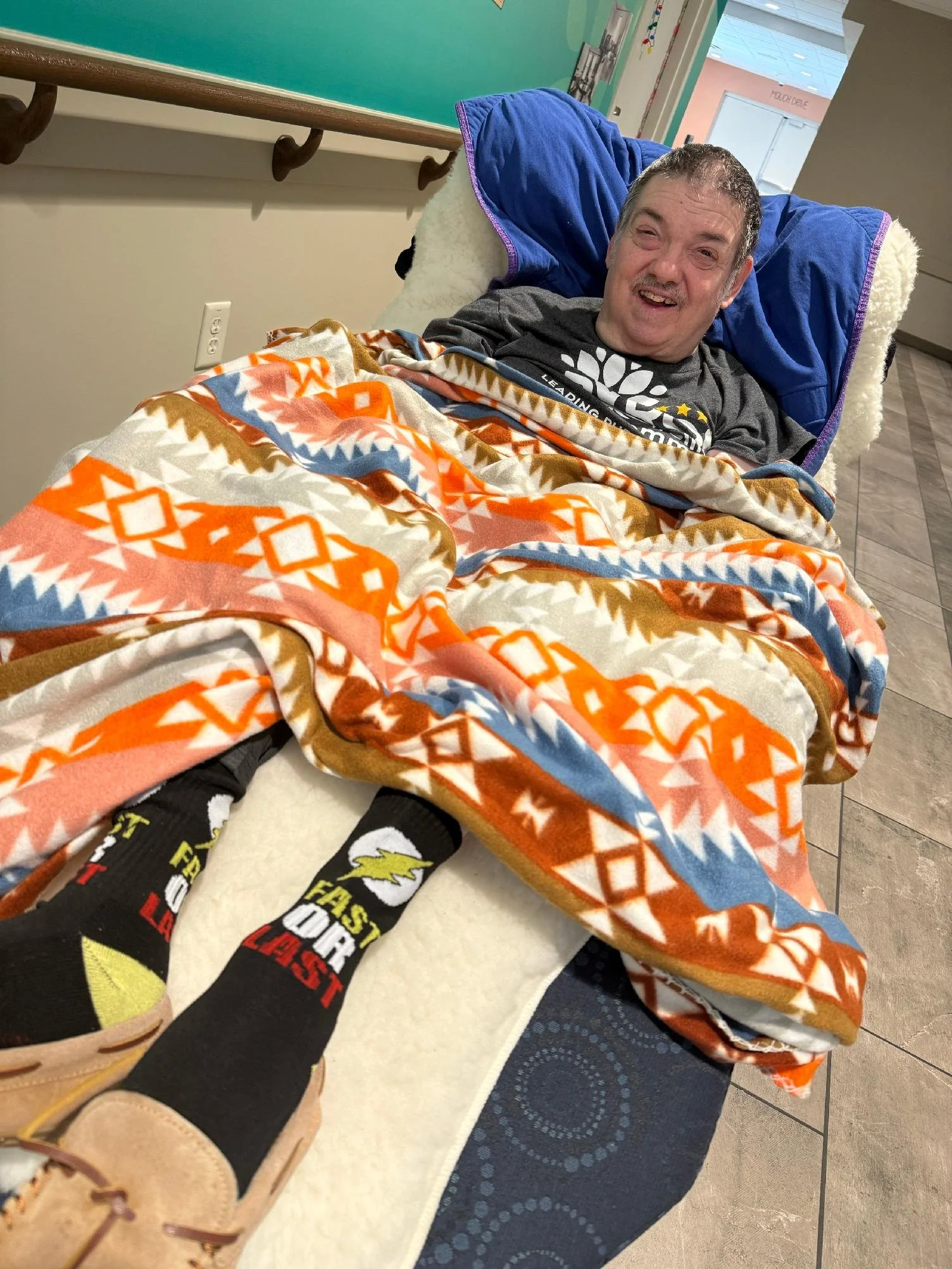 A man with short gray hair sitting in a hospital bed, smiling. He is covered with a colorful blanket and wearing black socks with colorful graphics, including a lightning bolt and text.