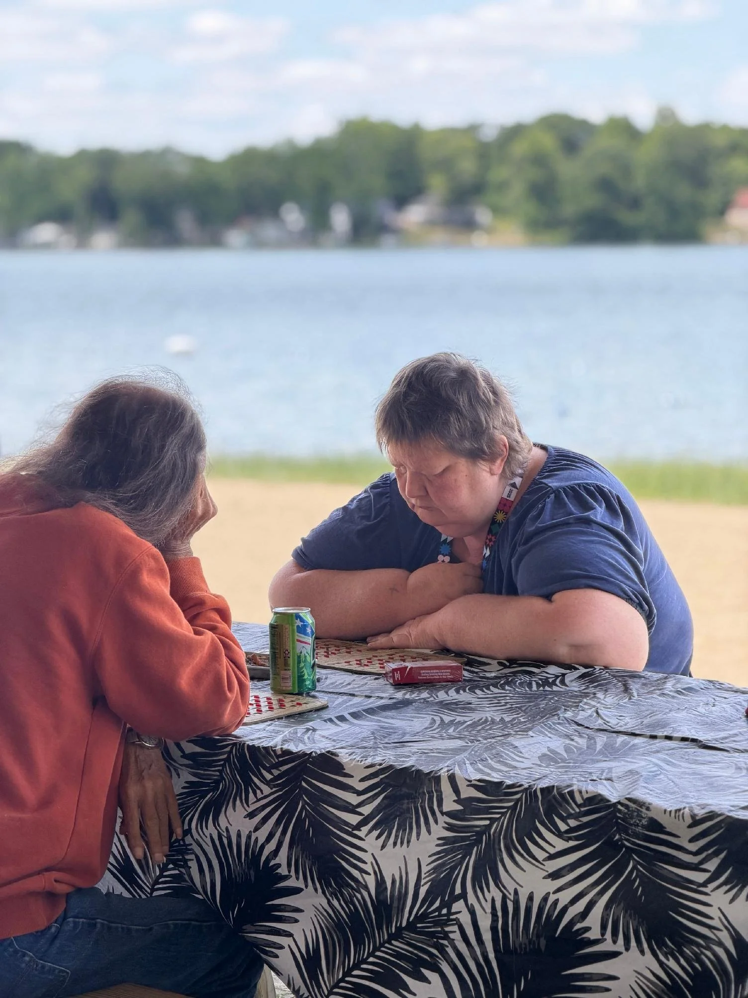 Two women sitting at a table outdoors near a lake, with trees and houses in the background. The woman on the left has gray hair and is wearing an orange hoodie, while the woman on the right has short hair, is wearing a navy shirt, and appears to be f