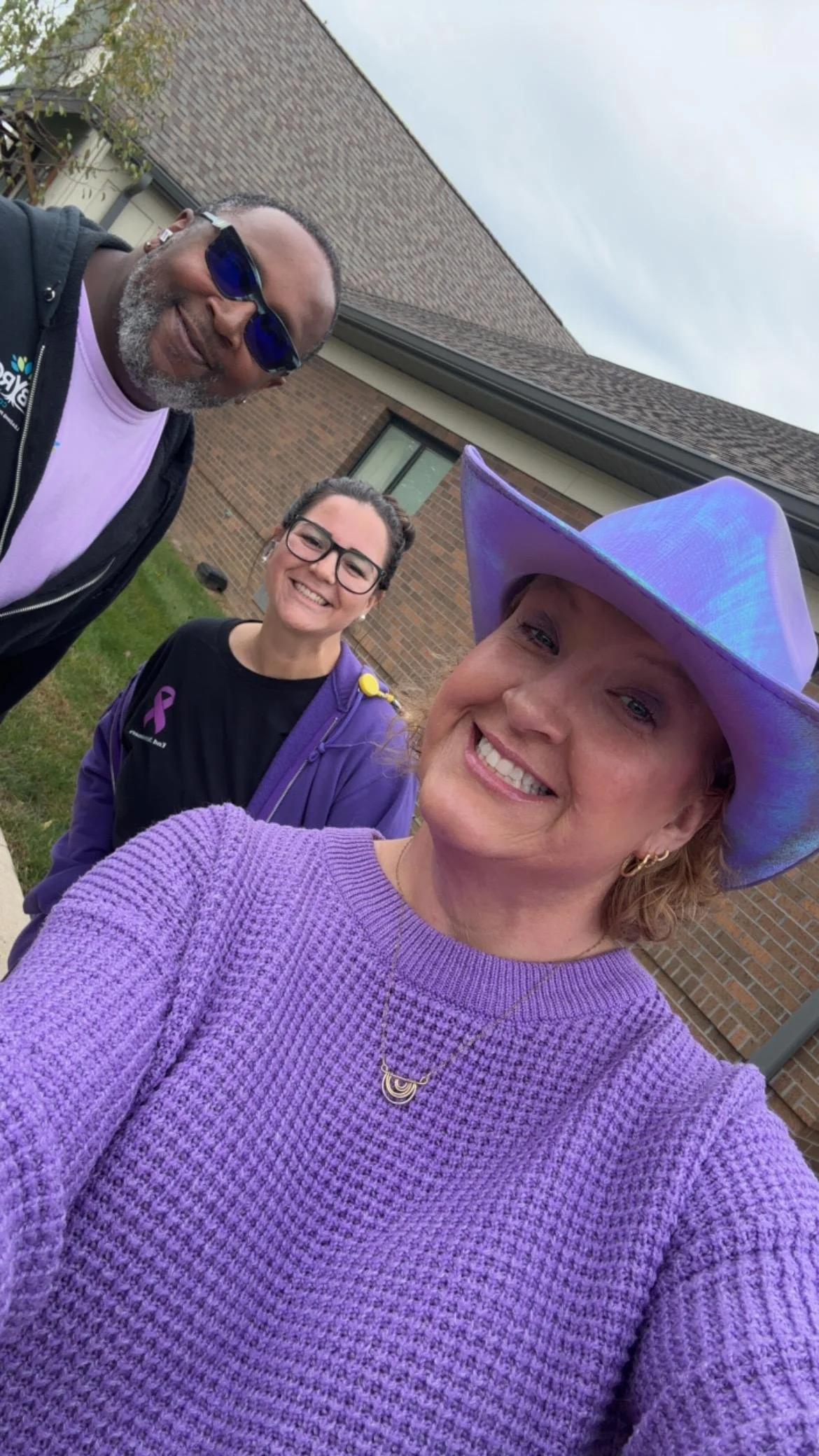 Three women taking a selfie outdoors in front of a brick building. The woman in the foreground is wearing a purple sweater, gold earrings, and a large purple hat. The woman in the middle is wearing glasses and a purple jacket, and the woman on the le