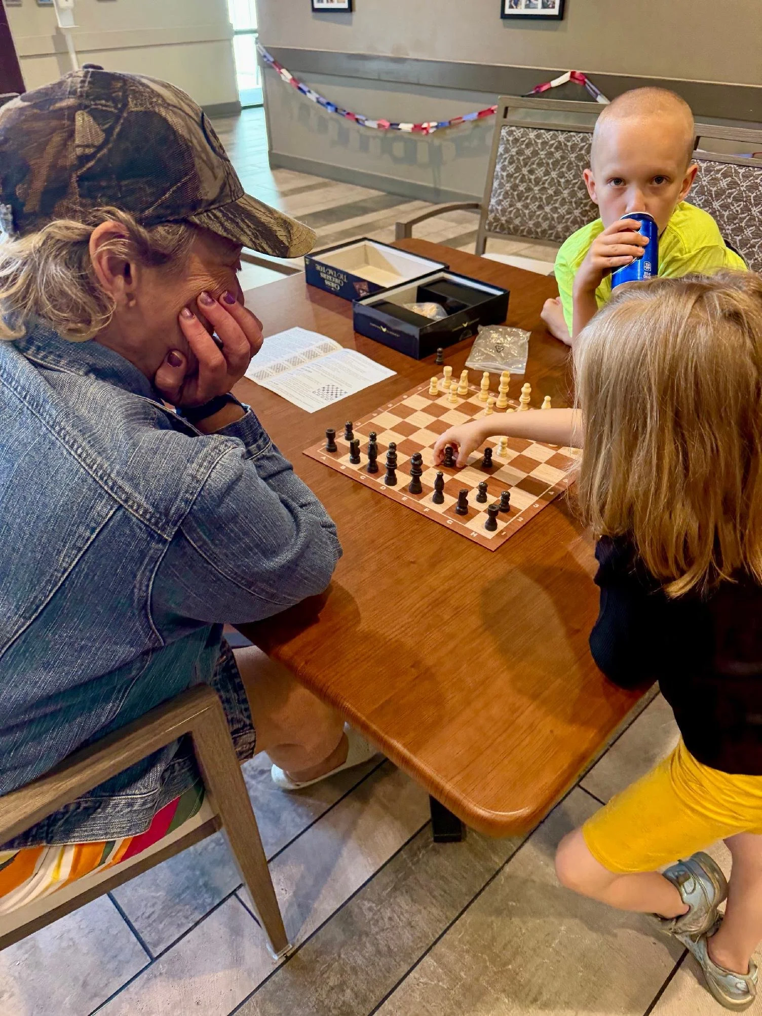 A woman and two children sitting at a wooden table playing chess in a cafe. The woman is wearing a camouflage cap and denim jacket, and the children are wearing bright yellow shorts and shirt, with one boy drinking from a can.