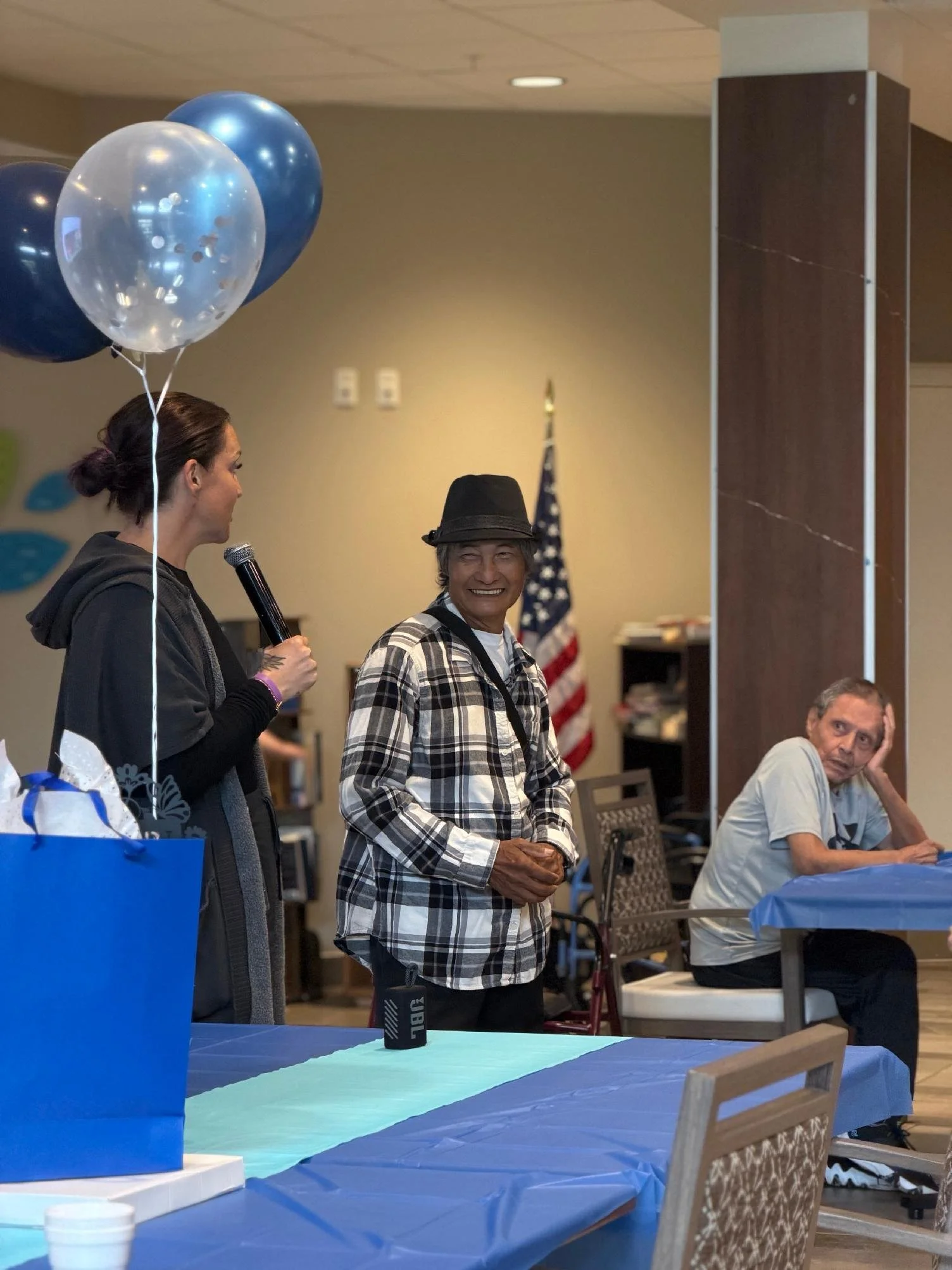 A woman is giving a speech with a microphone, holding some balloons, while smiling at a man who is standing nearby. The man is wearing a plaid shirt and a black hat, and is smiling back. In the background, another man is sitting at a table, resting h