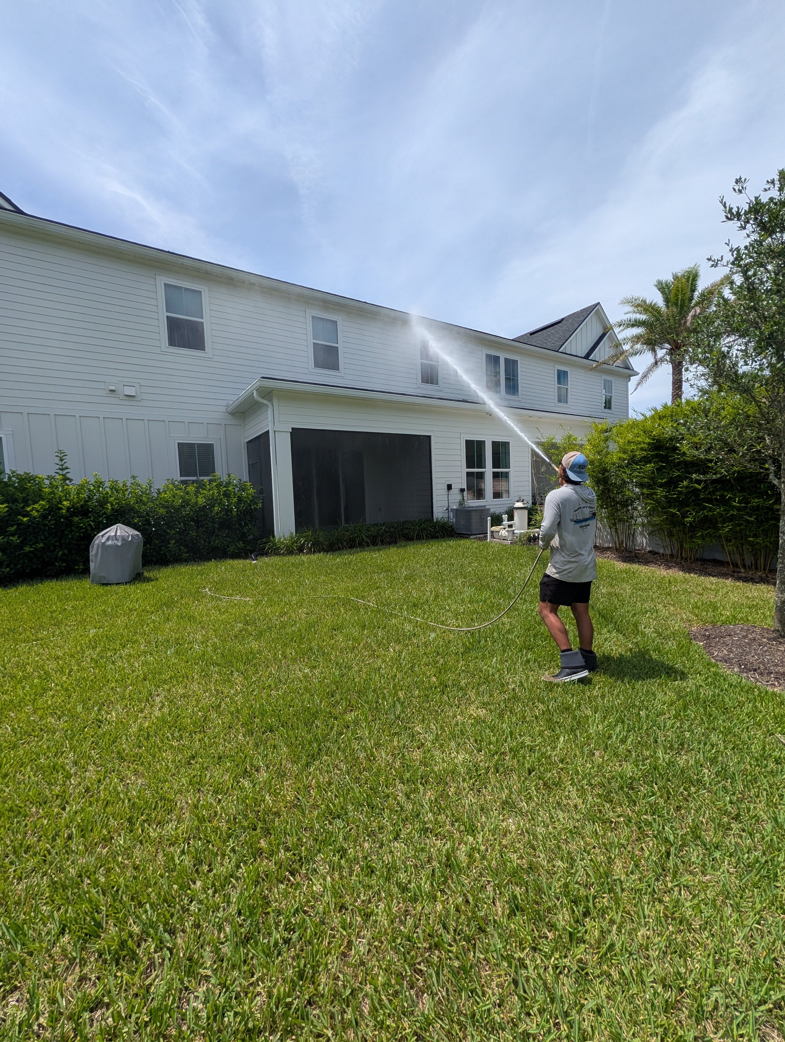 A man wearing a hat and shorts sprays water on a green lawn in a backyard. There are bushes, a covered object, and a house with multiple windows in the background.