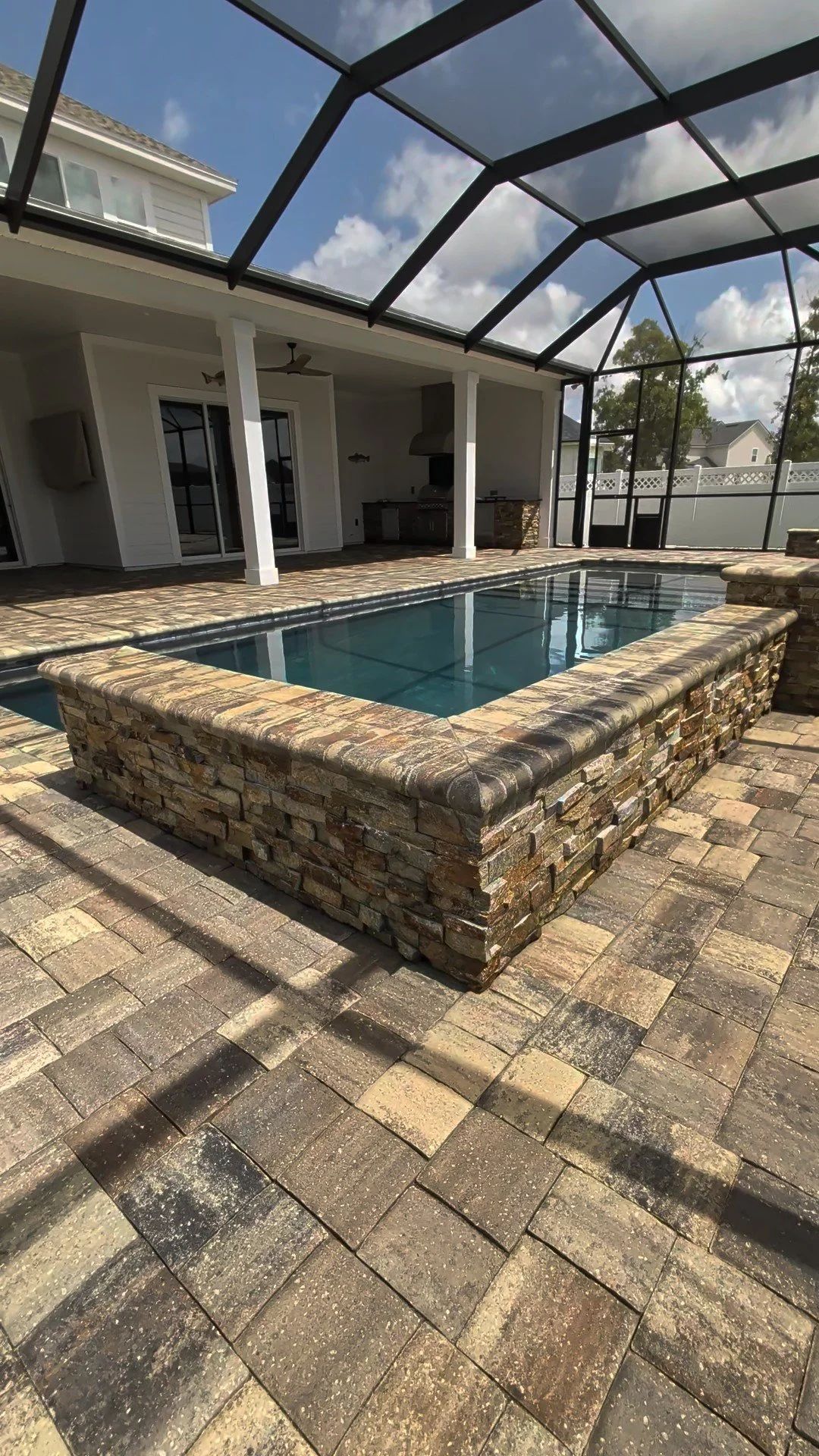 Backyard patio with a small rectangular pool surrounded by stone tiles and a screened enclosure, with a covered house patio featuring a ceiling fan and outdoor kitchen area.