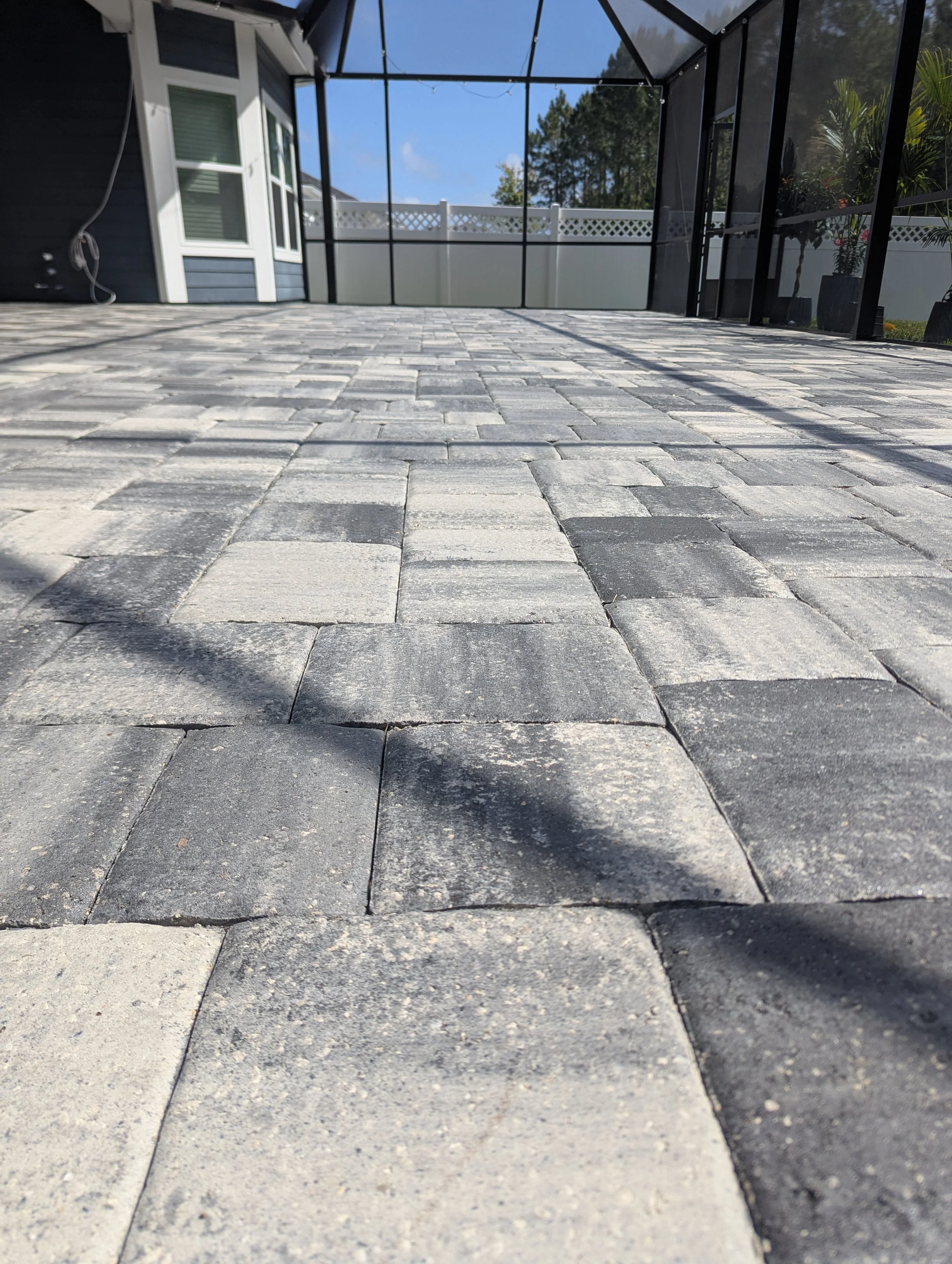 View of a backyard patio with stone pavers, screened enclosure, and a white fenced yard with trees in the background.