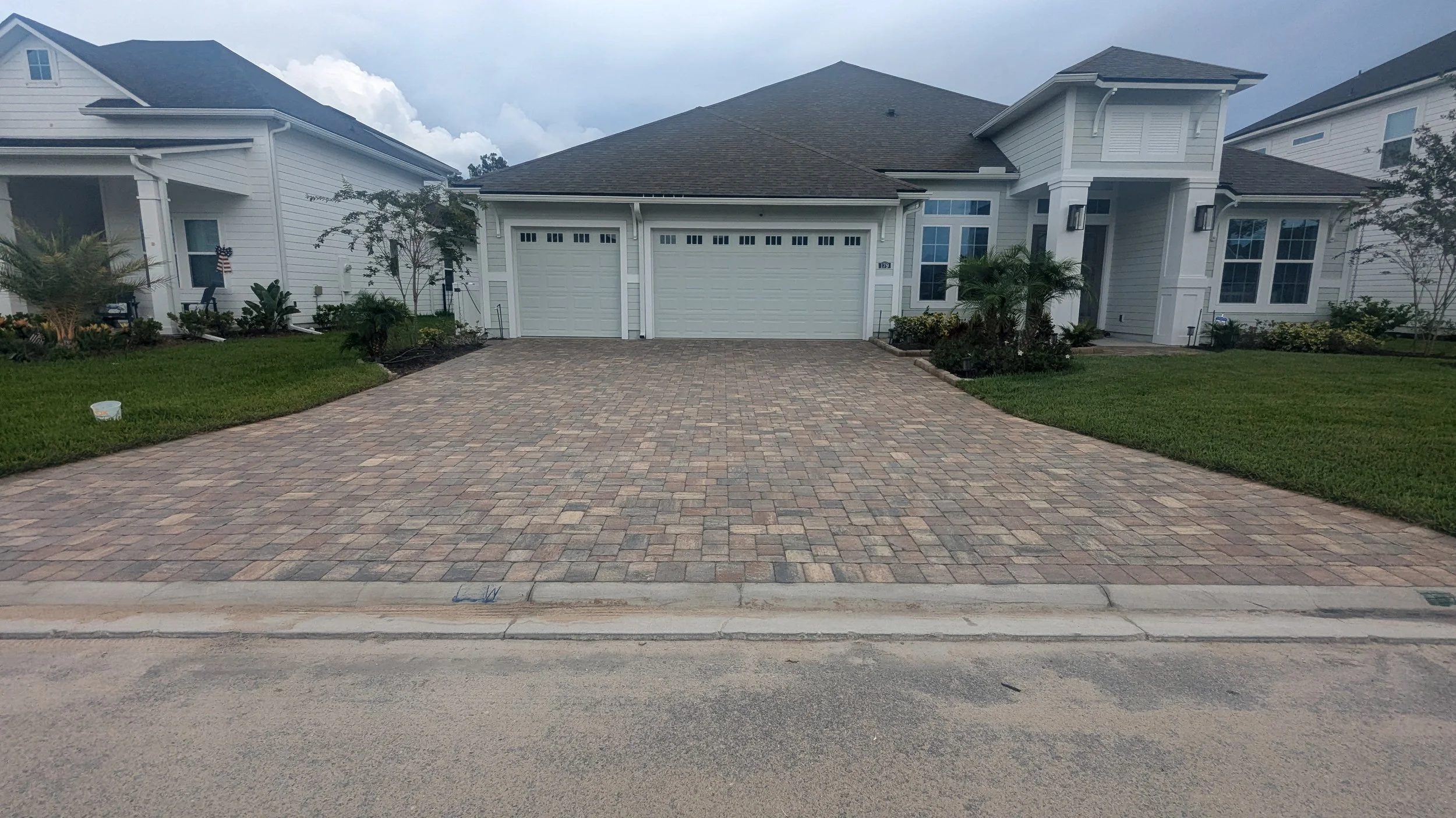 A newly paved brick driveway in front of a modern white house with a two-car garage, green lawn, small trees, and landscaping, under a cloudy sky.