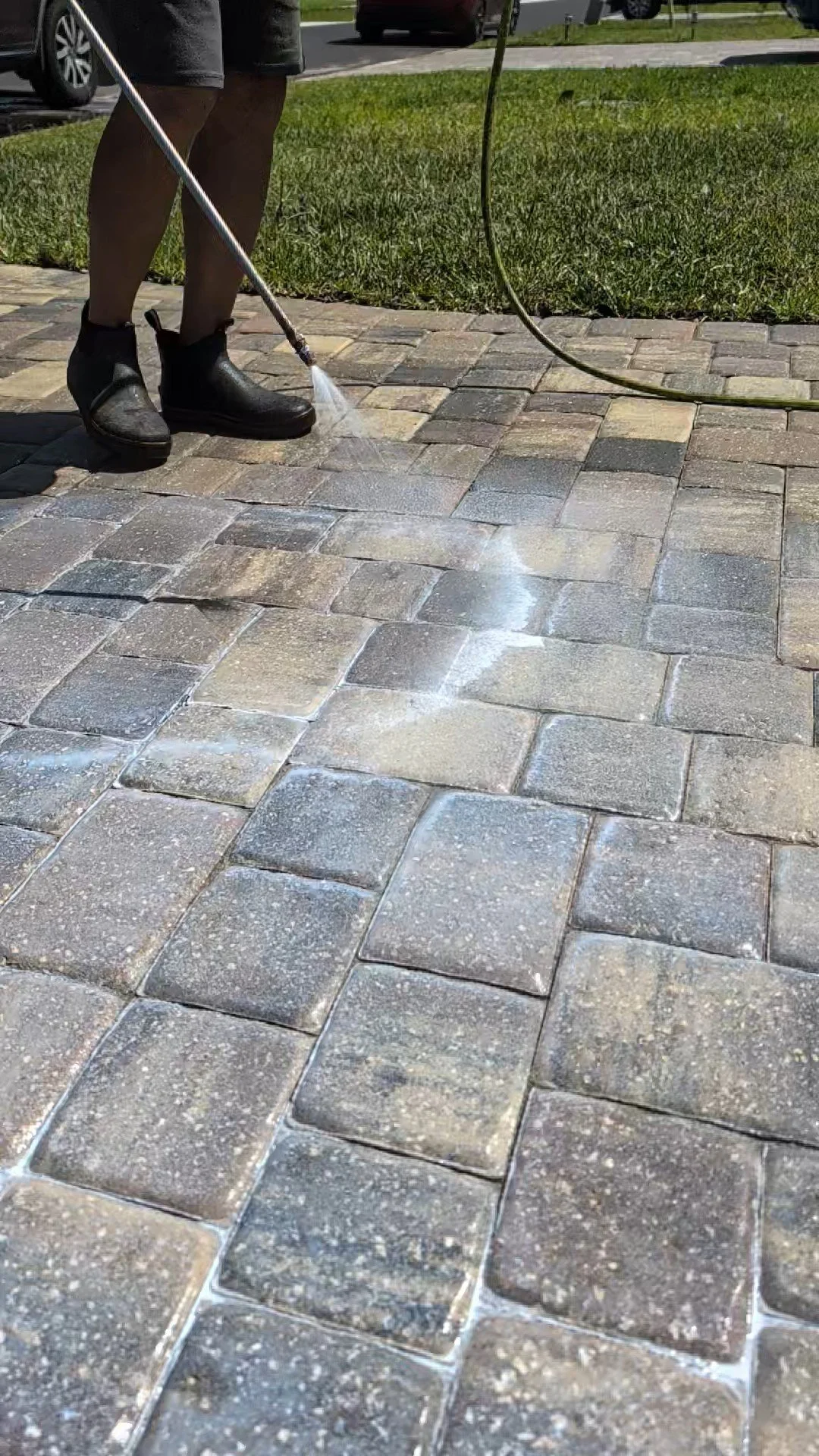 Person pressure washing a brick patio with water and cleaning solution, wearing black boots, black shorts, and holding a nozzle connected to a hose.
