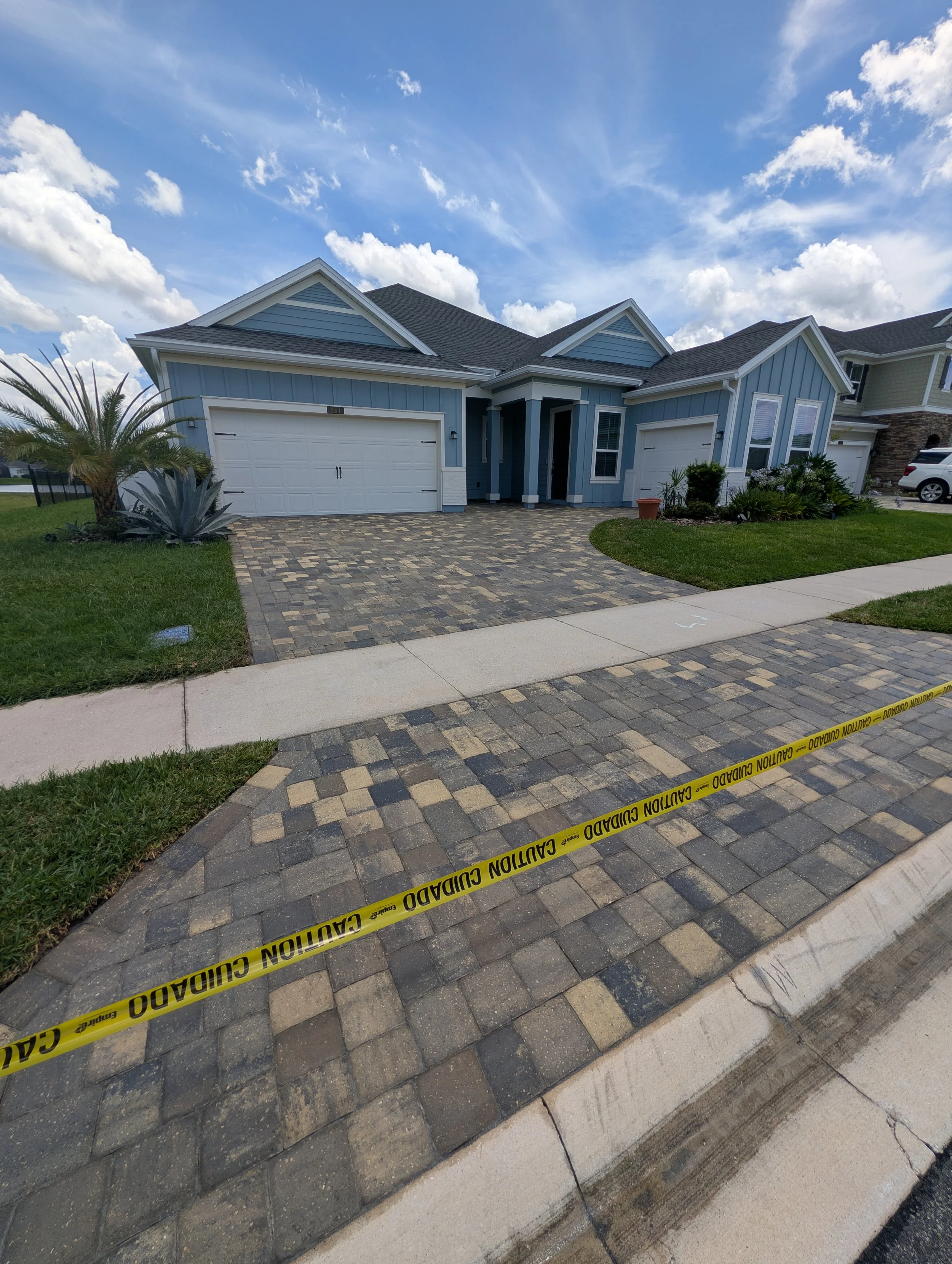 A two-story house with a light blue exterior, white garage doors, and a small front garden. The house is surrounded by a brick driveway and sidewalk. Yellow caution tape with 'Caution Guard' written on it is across the front, indicating a restricted area.