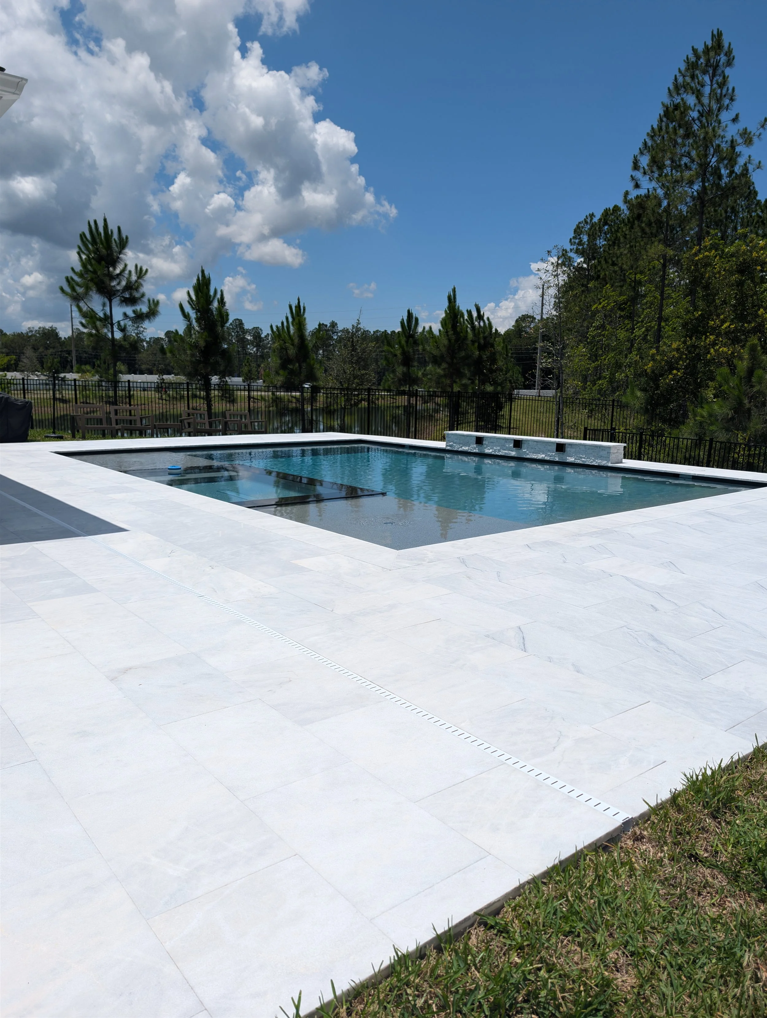 An outdoor swimming pool with a white tiled deck surrounded by a black metal fence. In the background, there are tall pine trees, a grassy area, and a partly cloudy blue sky.