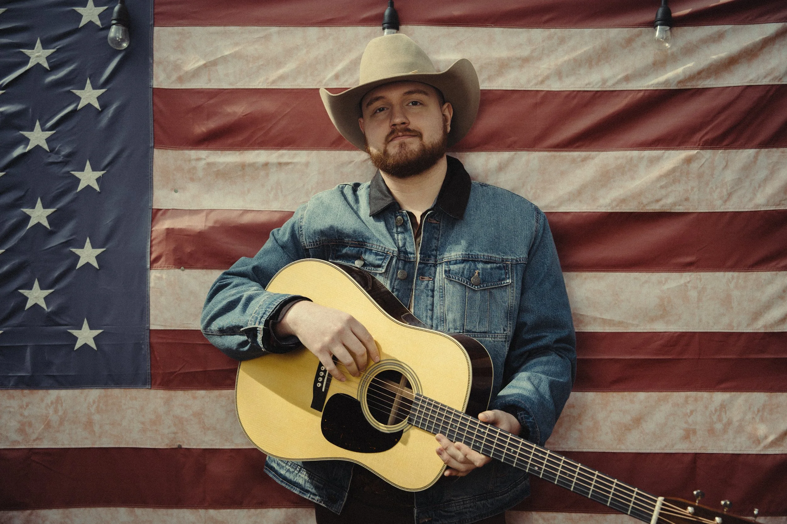 A man with a beard wearing a cowboy hat and denim jacket is holding an acoustic guitar, standing in front of a large American flag backdrop.