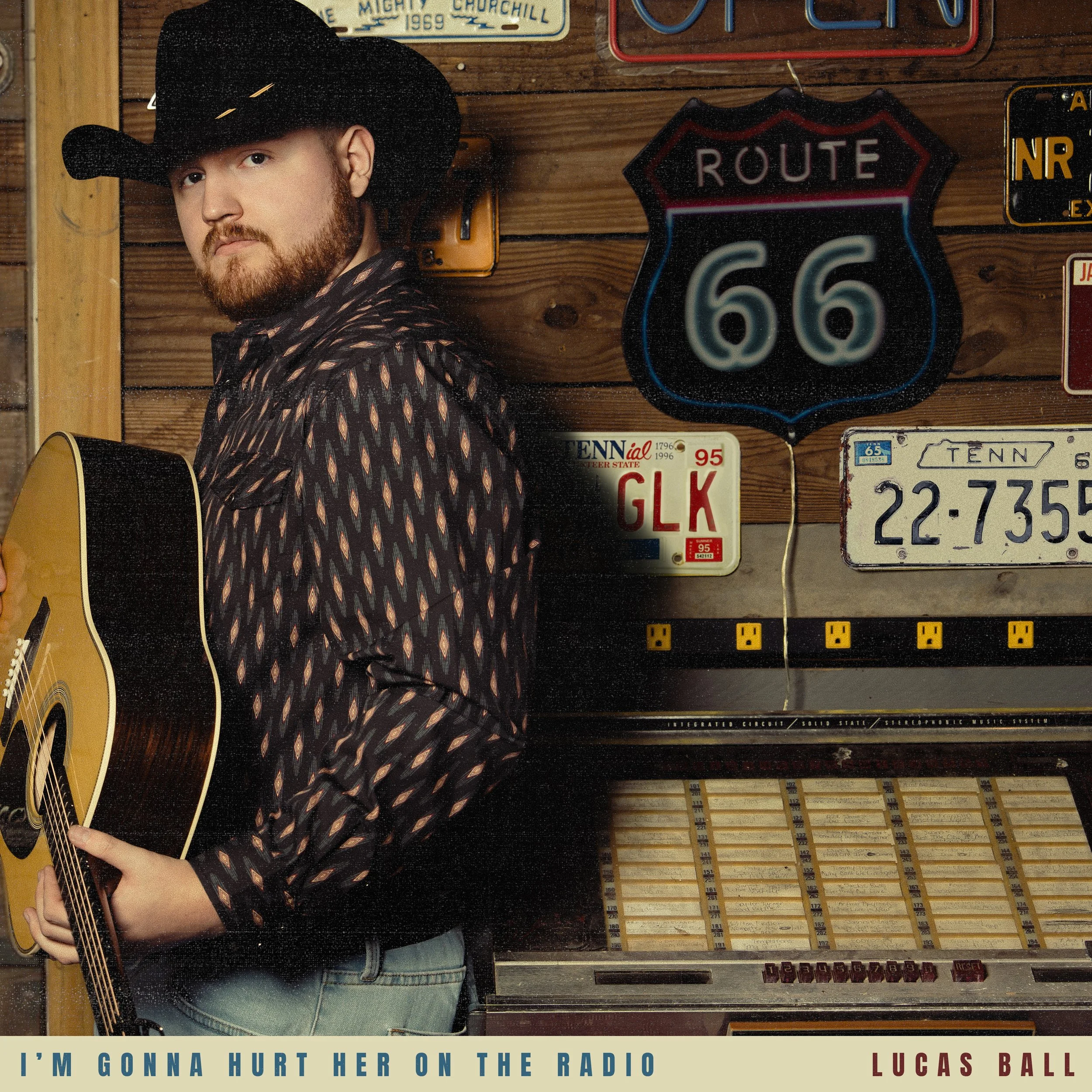 A man with a beard wearing a black cowboy hat and patterned shirt, playing an acoustic guitar, standing in front of a wall decorated with vintage road signs and license plates, including a Route 66 sign and Tennessee license plates. There is a caption at the bottom that reads, 'I'M GONNA HURT HER ON THE RADIO LUCA'S BALL.'