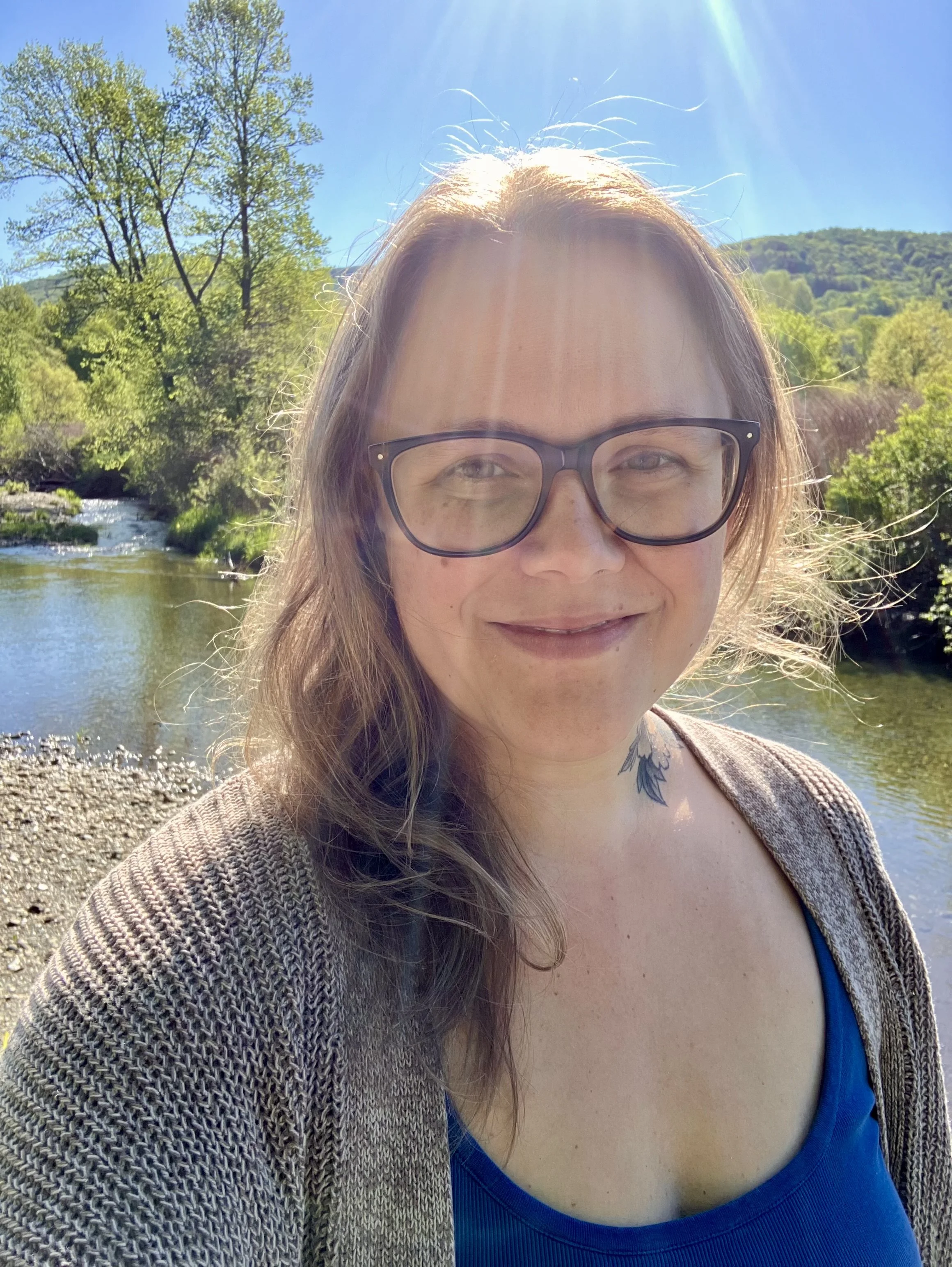 A woman with brown hair and glasses smiling outdoors near a river, with trees and hills in the background on a sunny day.