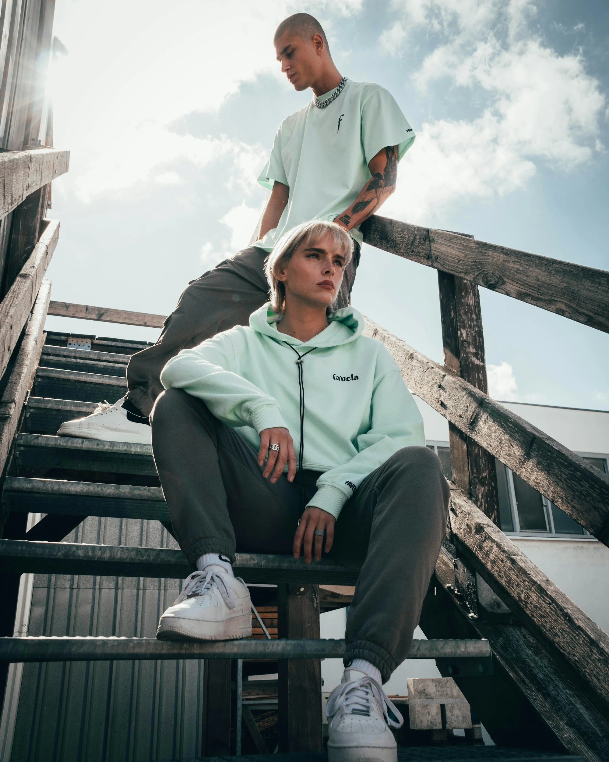 Two people sitting on outdoor staircase with a man standing above a woman. Both are dressed casually, with the woman in a light green hoodie and the man in a white t-shirt. The woman has short blond hair and the man is bald with tattoos on his arm. T