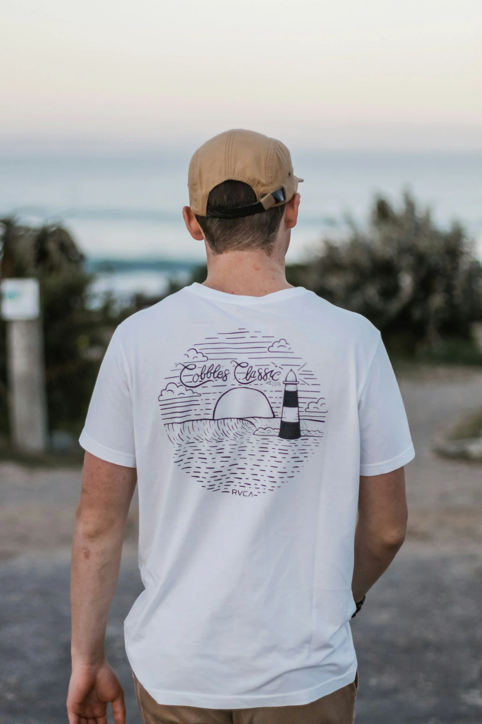 A person wearing a light brown baseball cap and a white T-shirt standing outdoors with the ocean and some trees in the background, seen from behind.
