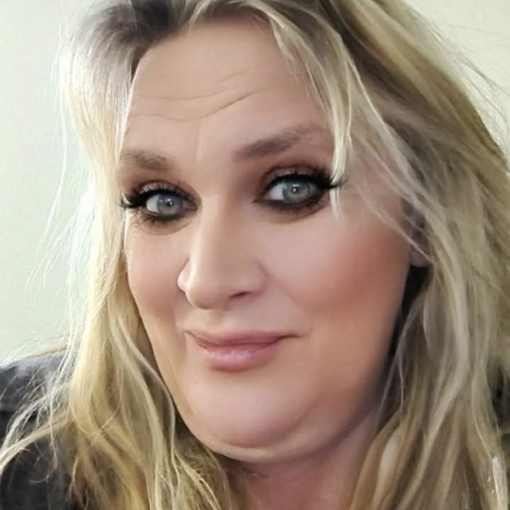 Close-up of a woman with blonde hair and blue eyes, wearing makeup, smiling gently.