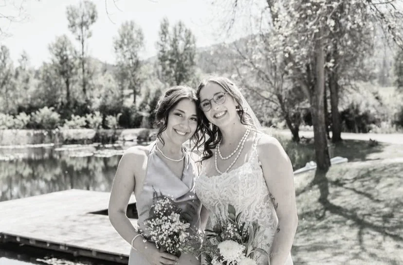 Two women dressed in formal attire holding bouquets of flowers, standing outdoors near a lake with trees in the background.
