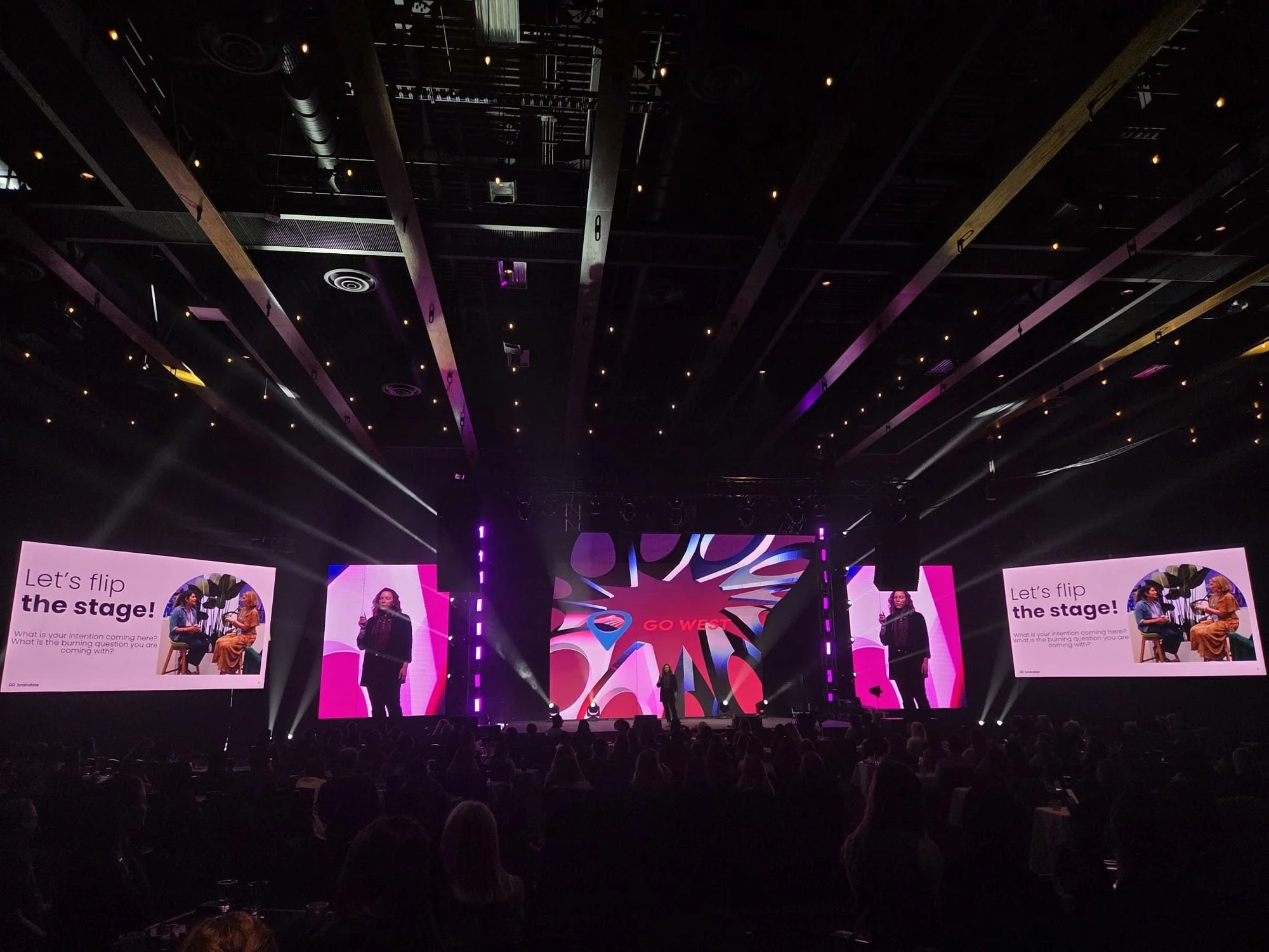 Christine Renaud on stage. A large indoor stage at a conference with two screens on either side displaying the words "Let's flip the stage" and images of people. The stage has bright lights and a colorful abstract background on a central screen.