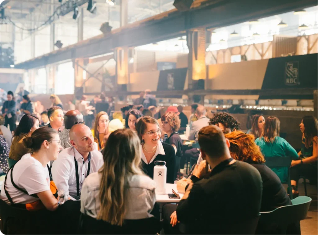 Braidate Networking. People gathered around tables in a large, industrial-style event space with high ceilings and natural light, engaging in conversations.