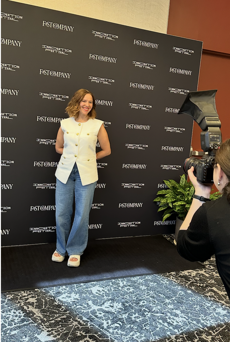 Christine Renaud standing on a black platform with a backdrop reading 'Fast Company' and 'Innovation Festival'. She is smiling, wearing a white sleeveless vest, blue jeans, and heels, while a photographer takes her picture.