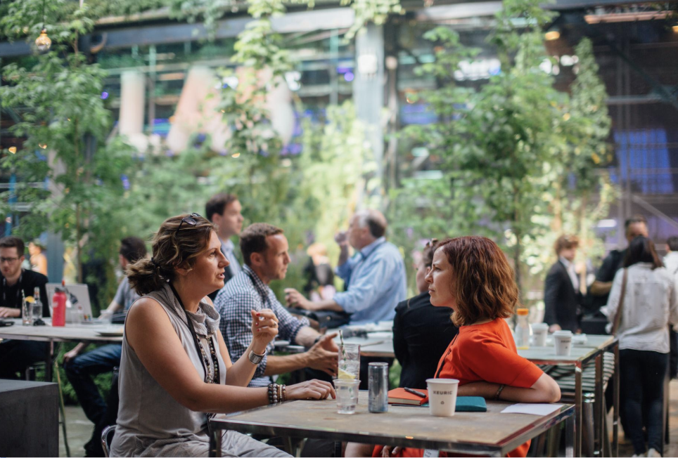 Braindate networking. People sitting and talking at tables in a lush, green indoor space resembling a garden or greenhouse, with multiple groups engaging in conversations.