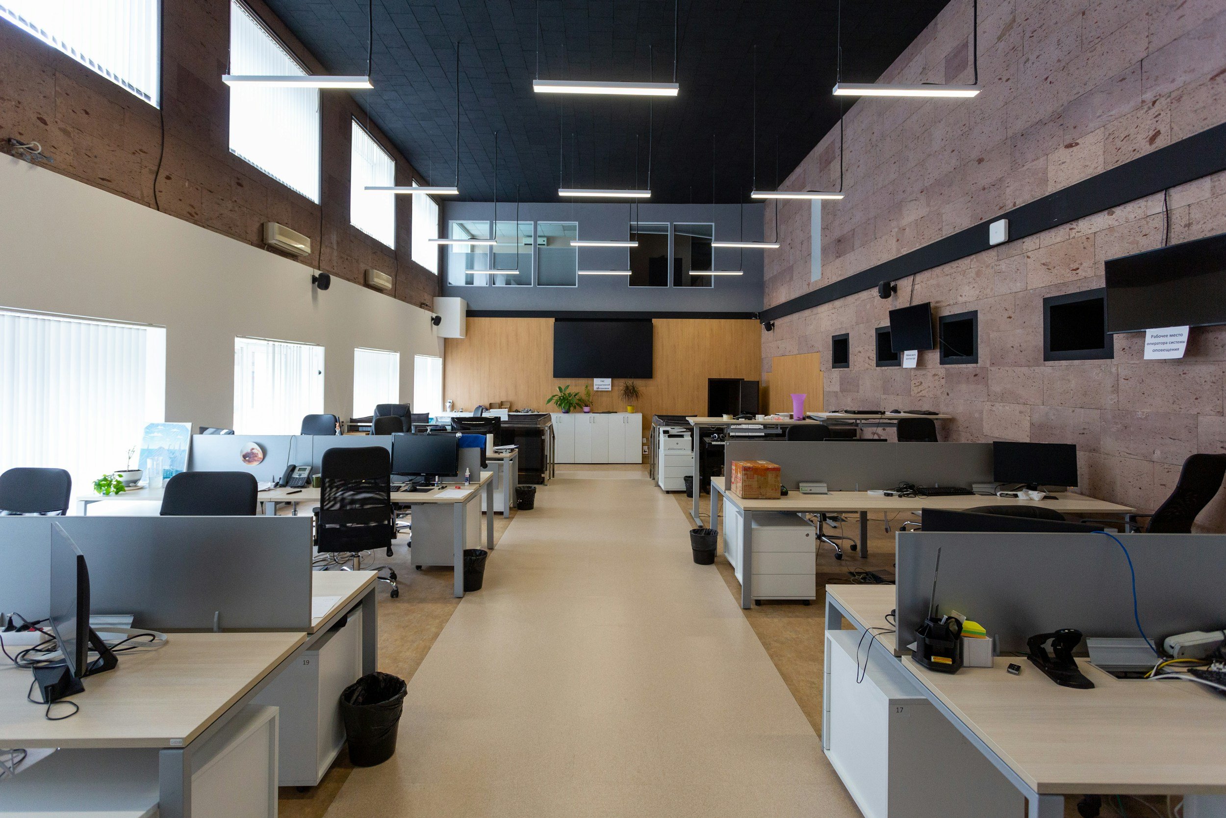 Empty office with desks, chairs, and computer monitors, beige walls, large windows with blinds, and ceiling lights.