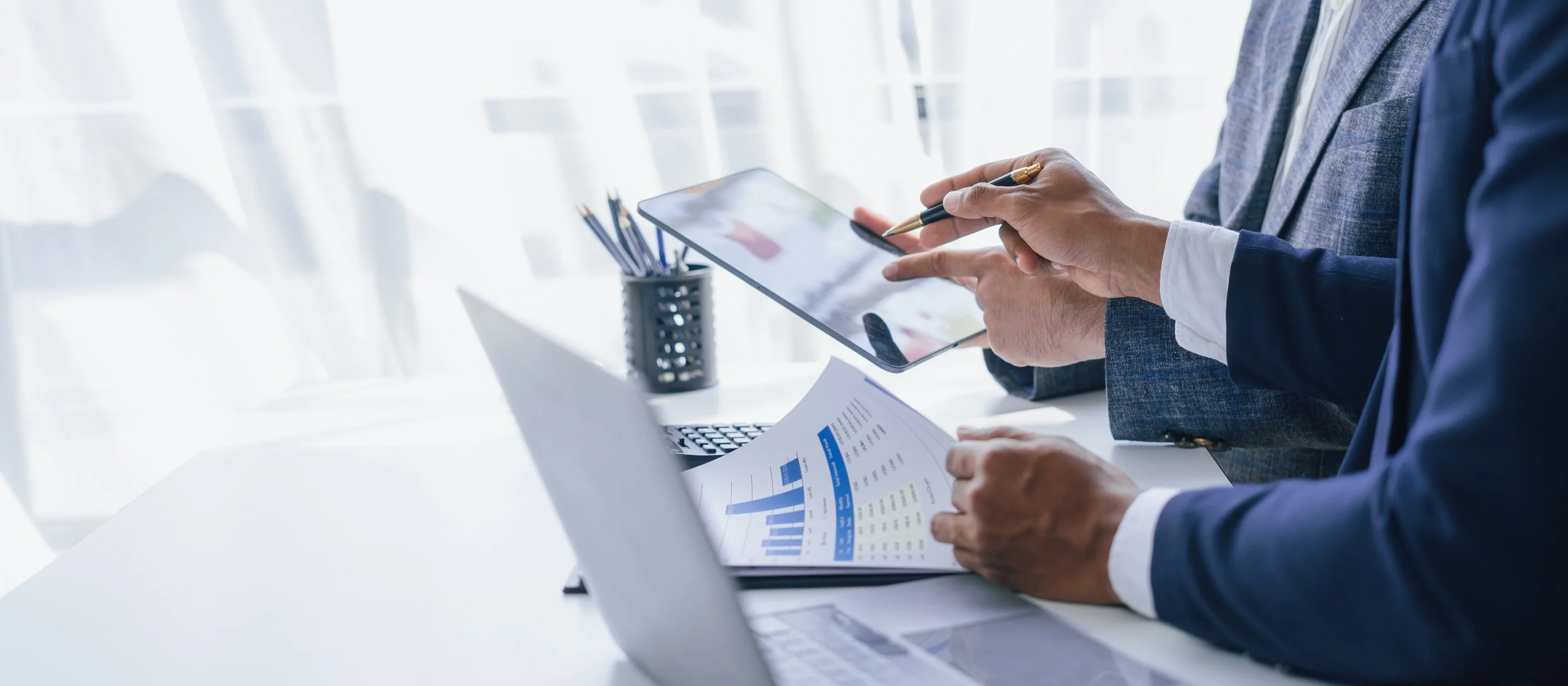 Business professionals working at a desk, one person holding a tablet and a pen, another reviewing financial reports with charts and graphs.