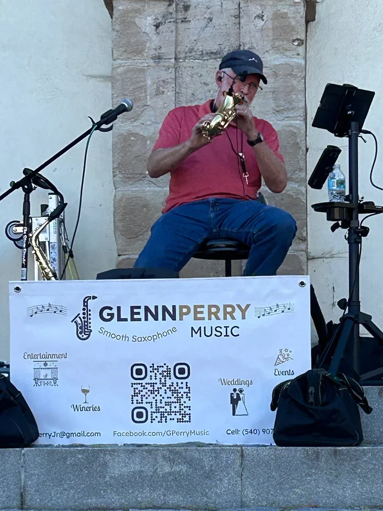A man in a red shirt, blue jeans, and a black cap playing a alto saxophone on a small outdoor stage. There are music equipment and a banner with contact information for Glenn Perry, a smooth saxophone music performer.