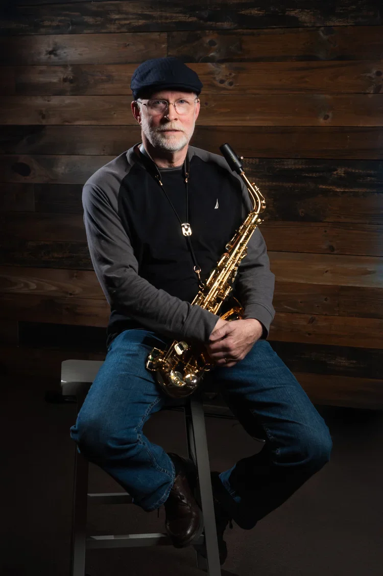 A man with glasses and a beard, wearing a black beret and gray jacket, sitting on a stool holding a saxophone against a dark wooden wall background.