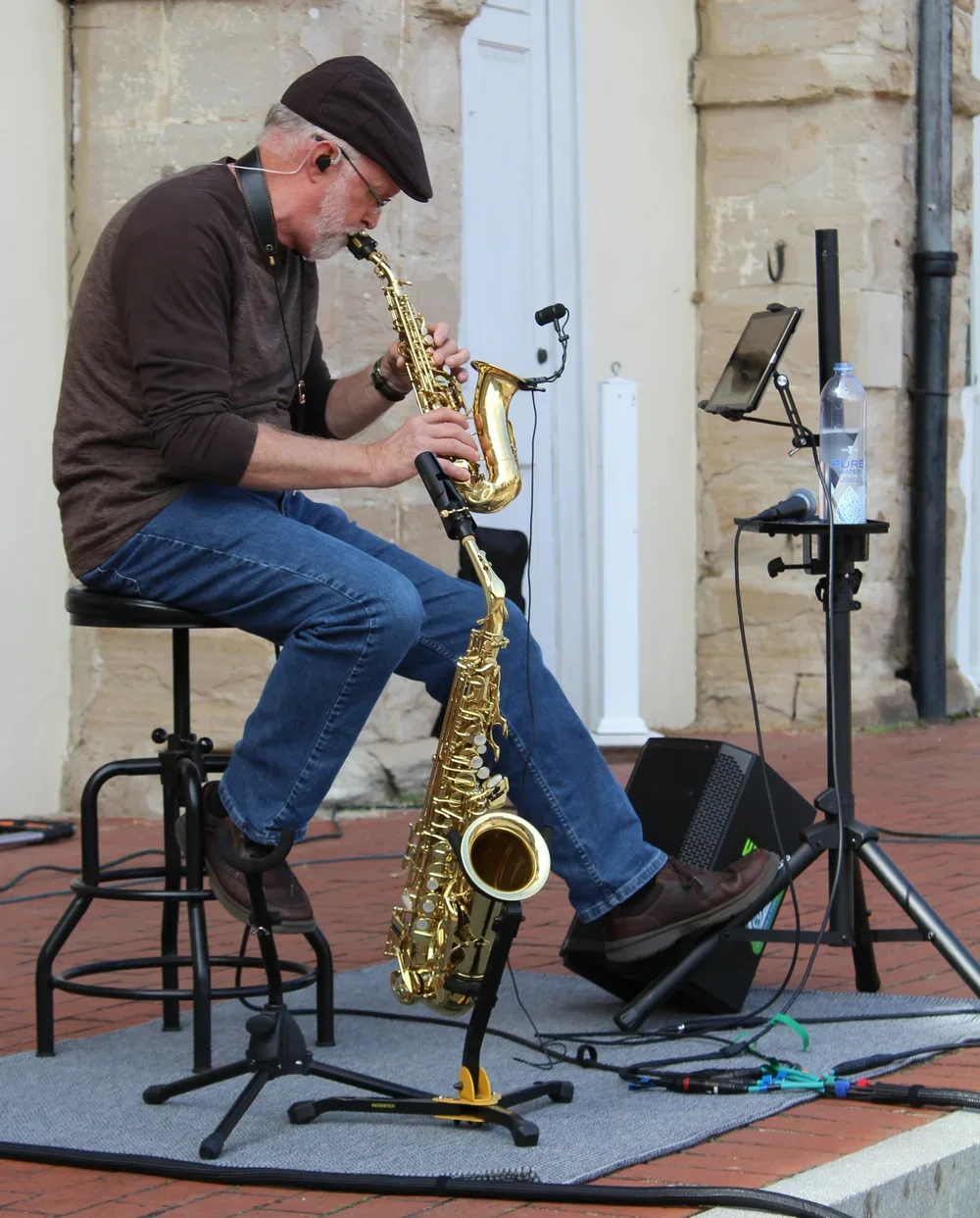 A man sitting on a stool playing a soprano saxophone with an alto saxophone resting on the ground nearby, set up outdoors with audio equipment including a microphone, speaker, and tablet for a performance.