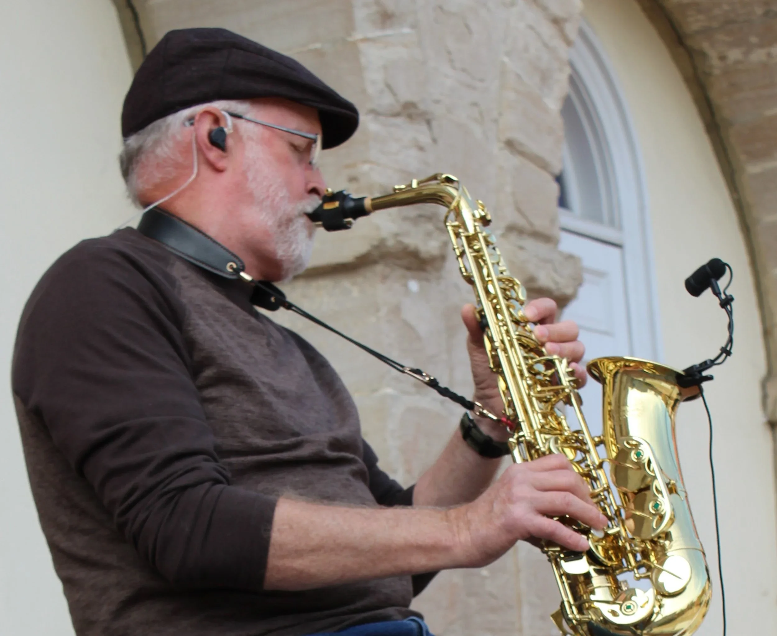 An older man with gray hair, glasses, and a dark flat cap playing a gold saxophone indoors, with a stone wall and window in the background.