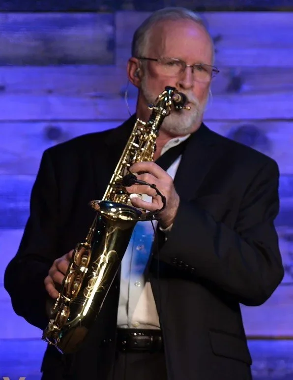 A man with glasses playing a saxophone on stage with a wooden backdrop.
