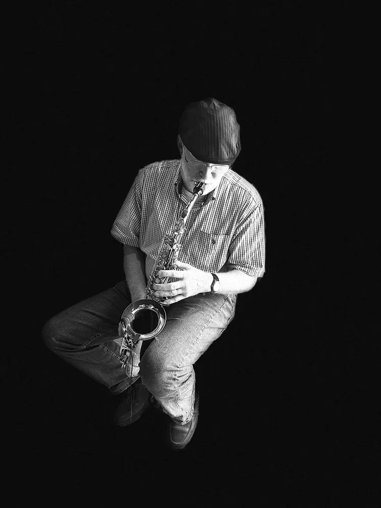 A man wearing a cap, glasses, and a checked shirt playing an alto saxophone against a black background.