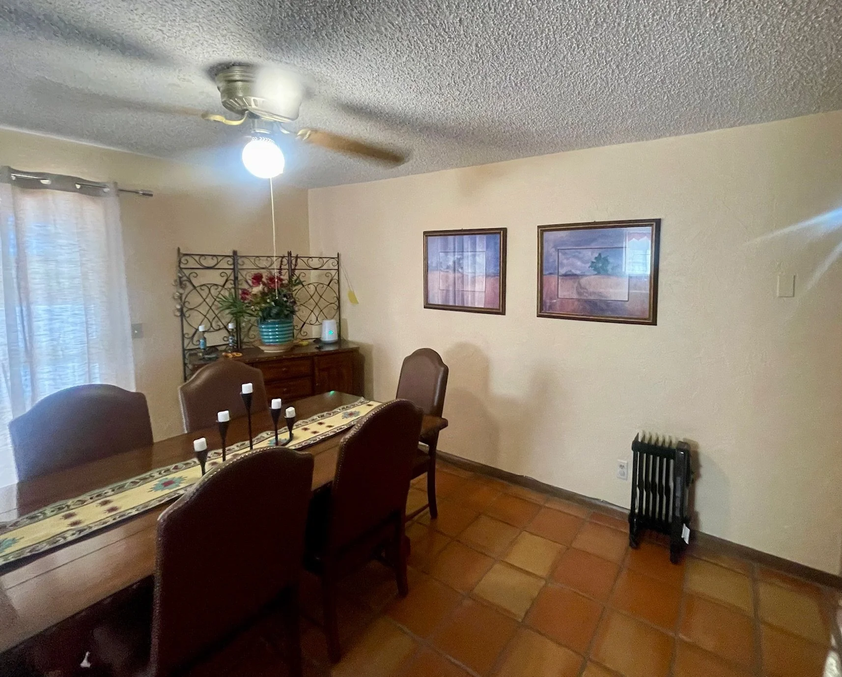 A dining room with a wooden table and six chairs, a ceiling fan with a light, a wall with two framed landscape paintings, a window with curtains, a sideboard with a plant and a white device, and a radiator.